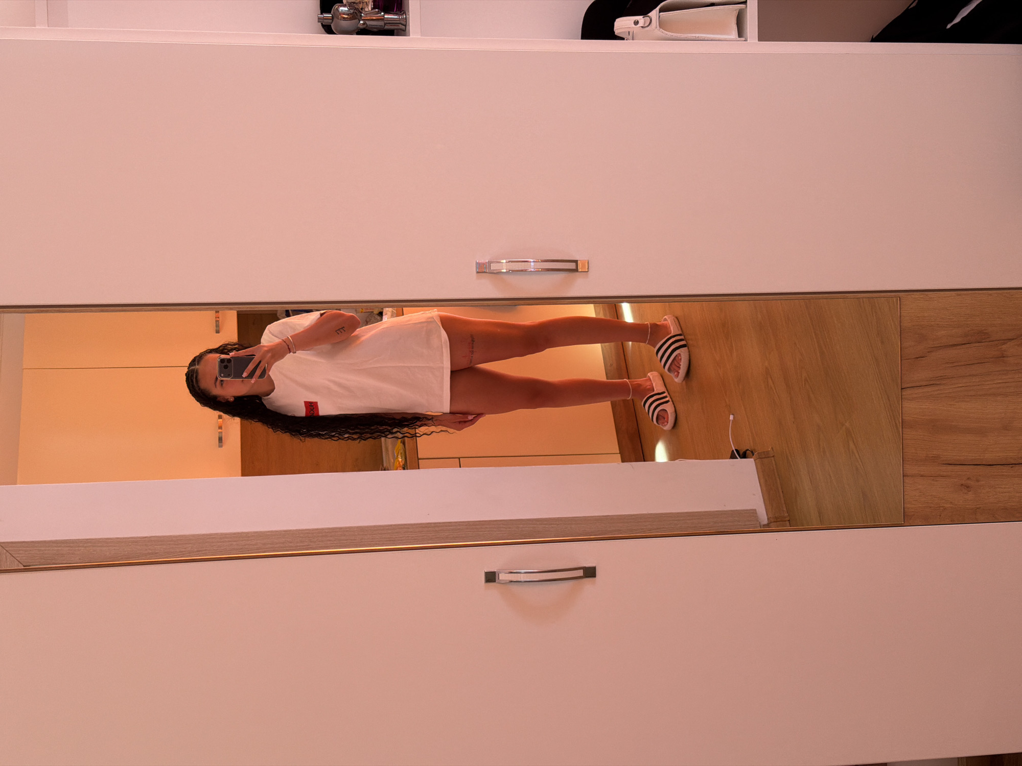 The image shows a young woman with long, dark, curly hair taking a mirror selfie in what appears to be a bedroom or dressing area. She is wearing a white t-shirt and white slides with black stripes. Her reflection is visible in a large mirror that spans the length of what seems to be a wardrobe. The background behind her reflection shows a kitchen area with orange-toned cabinets. The wardrobe doors are closed, and the right side of the wardrobe has shelves with some items, including a white handbag. The floor is made of wood-patterned laminate. There is no indication of the specific location within Nesebar, Bulgaria.
