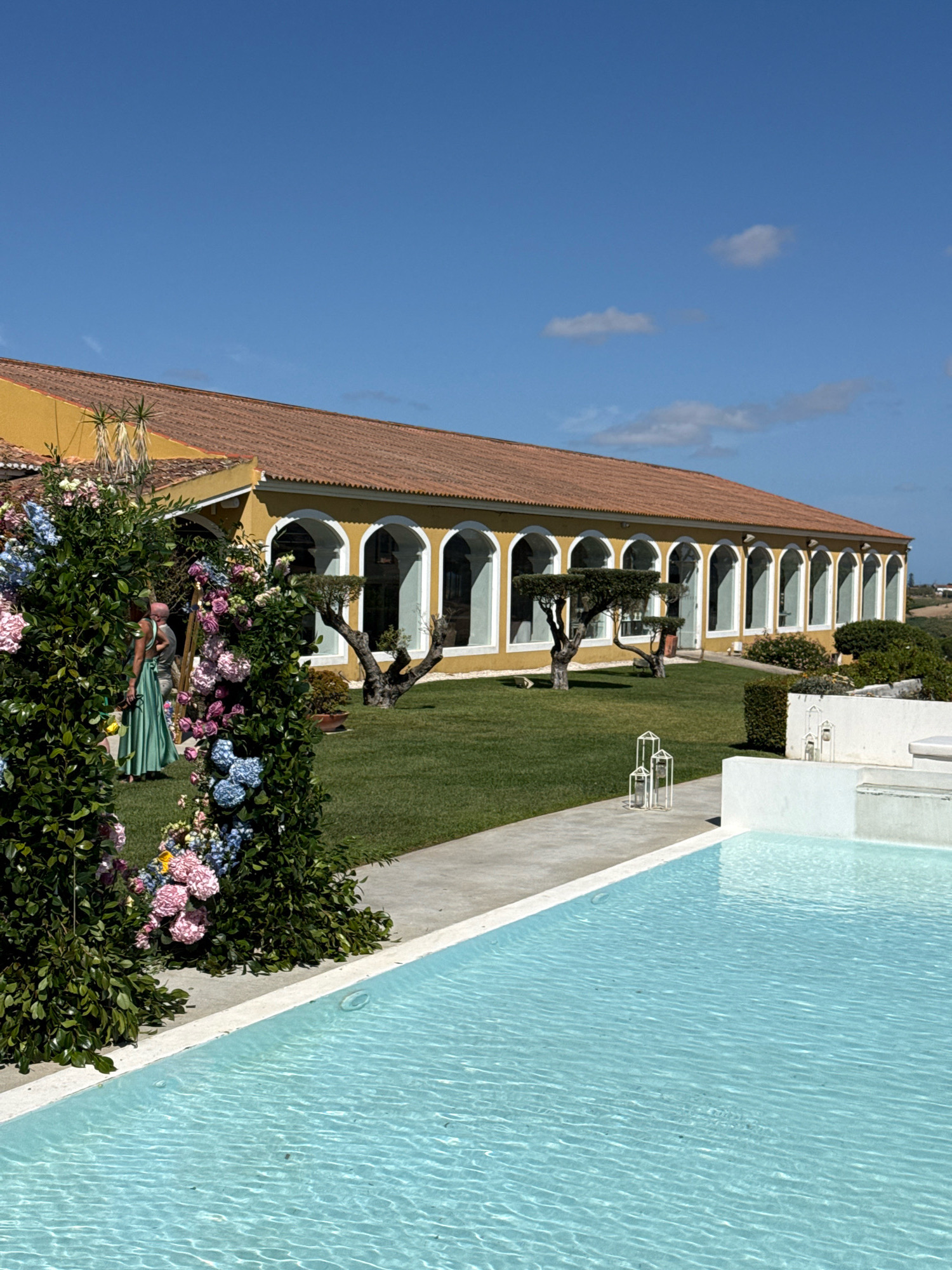 The image depicts a sunny day at a venue located in Mafra, Portugal. In the foreground, a bright blue swimming pool with visible ripples occupies the lower half of the frame. To the left of the pool, a walkway leads towards a building. Along the edge of the walkway and pool, there are lush green bushes adorned with clusters of pink and blue flowers, forming a decorative archway.
Further back, a long, yellow building with a terracotta tiled roof is visible. The building features a series of arched windows with white frames. Several trees with gnarled trunks are planted in the manicured green lawn in front of the building. Two individuals are present on the lawn; one person, wearing a long green dress, is partially obscured by the floral arrangement, and another person is standing near them. In the distance, a rolling landscape with some vegetation is visible under a clear blue sky with a few wispy clouds. White decorative lanterns are placed on the walkway near the pool.