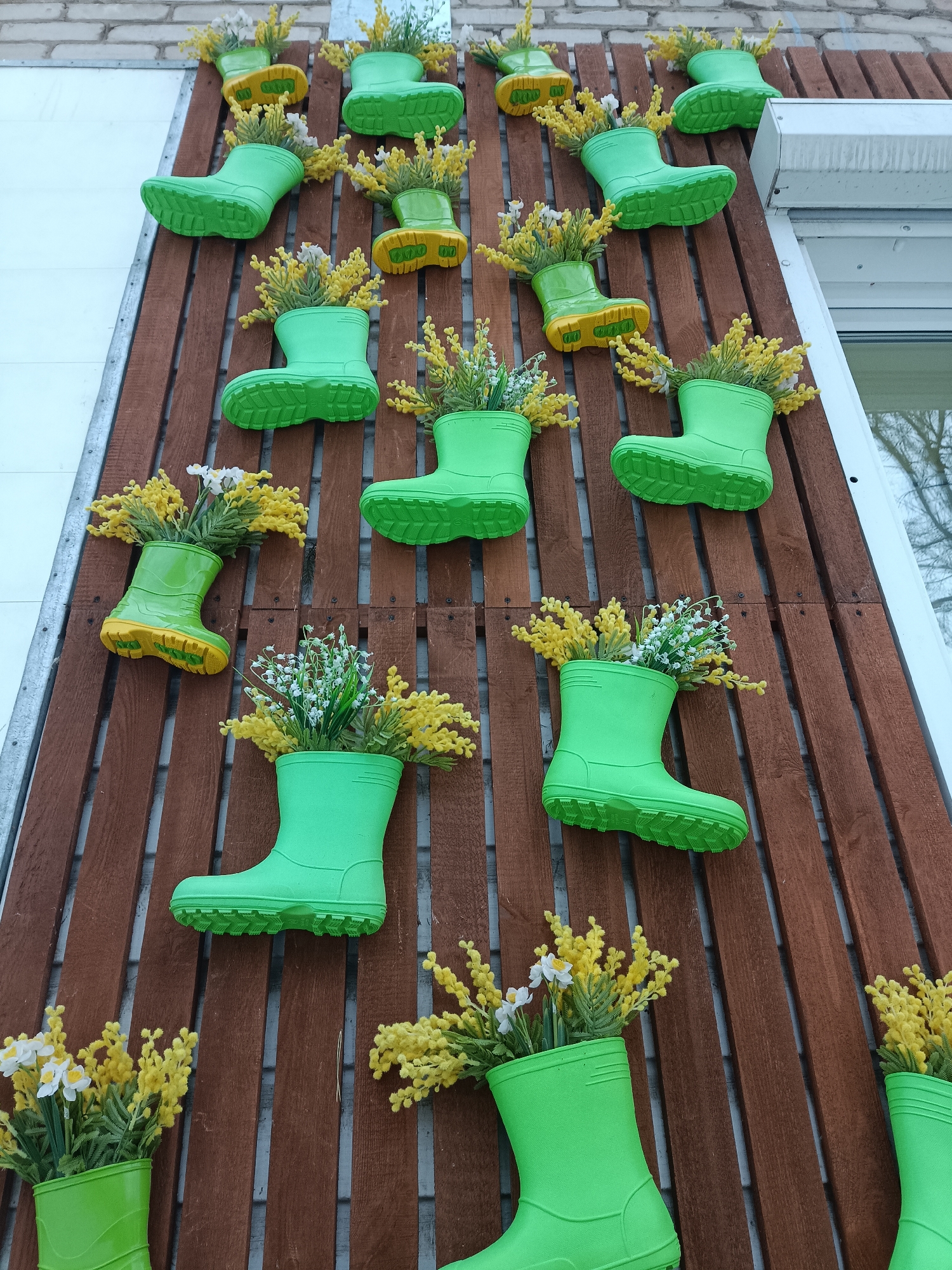 The image depicts an outdoor wall decoration located in Chaikovsky, Russia. Approximately 21-22 bright green rubber boots, some distinguished by yellow soles, are vertically affixed in a staggered pattern to a surface composed of horizontally arranged reddish-brown wooden slats. Each boot functions as a planter, holding clusters of artificial flowers; primarily yellow flowers resembling mimosa or acacia, with some boots also containing small white flowers akin to lily-of-the-valley or daffodils. The wooden slats are mounted onto a lighter-colored building facade, visible as brick or stucco. To the left, a segment of a white building wall with a vertical metallic grey beam is present. To the right, a white building fixture, possibly a window frame or vent, with a reflective glass pane showing faint tree reflections, is visible. No individuals are present in the scene.