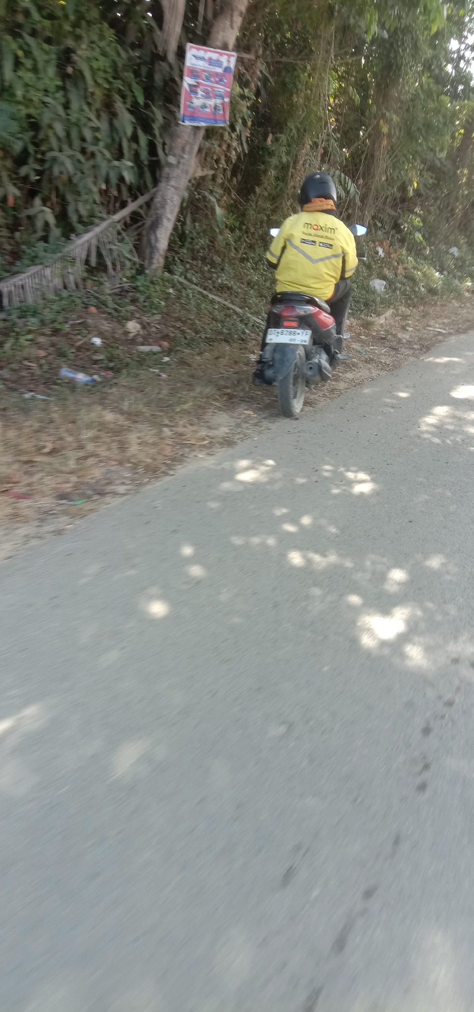 A person wearing a yellow jacket with "maxim" printed on it rides a scooter on a paved road in Kendari, Indonesia. The scooter's license plate is visible and reads "DT 5288 YF". The person is wearing a black helmet and a light brown scarf around their neck. The road is bordered by a dry, grassy area with some scattered trash. In the background, there are trees and foliage. A poster is affixed to a tree on the left side of the image. The sun is casting dappled shadows on the road, indicating it is daytime.