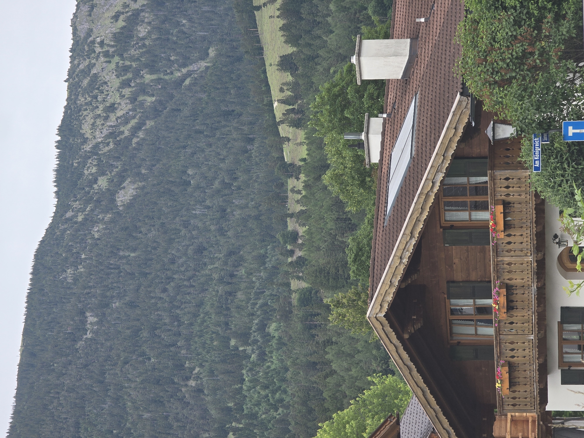 This image, when correctly oriented, presents a scenic view of a traditional Bavarian-style building set against a backdrop of a forested mountain. The building, situated in the foreground on the right, features a multi-peaked brown tiled roof with two chimneys and two skylights or solar panels. Its facade is constructed from dark wooden planks, including an ornate balcony with intricately carved railings adorned with window boxes filled with vibrant flowers. Part of a white-painted section of the building is visible on the far right.
Lush green trees and dense bushes surround the building. In the midground and background, a large mountain rises, densely covered with dark green coniferous trees. Scattered patches of lighter green foliage and exposed rocky areas are visible across the mountain slope. The sky is overcast or cloudy, indicating daytime with soft, diffused light.
No explicit human activity is taking place; the scene captures a static landscape and architectural view. A blue street sign is partially visible on the right, displaying the German text "Am Königreich" and a partially obscured word below, appearing to be "Stau." The architectural style, the mountainous terrain, and the German text on the sign collectively point to a location in the Bavarian Alps, consistent with Garmisch-Partenkirchen, Germany.