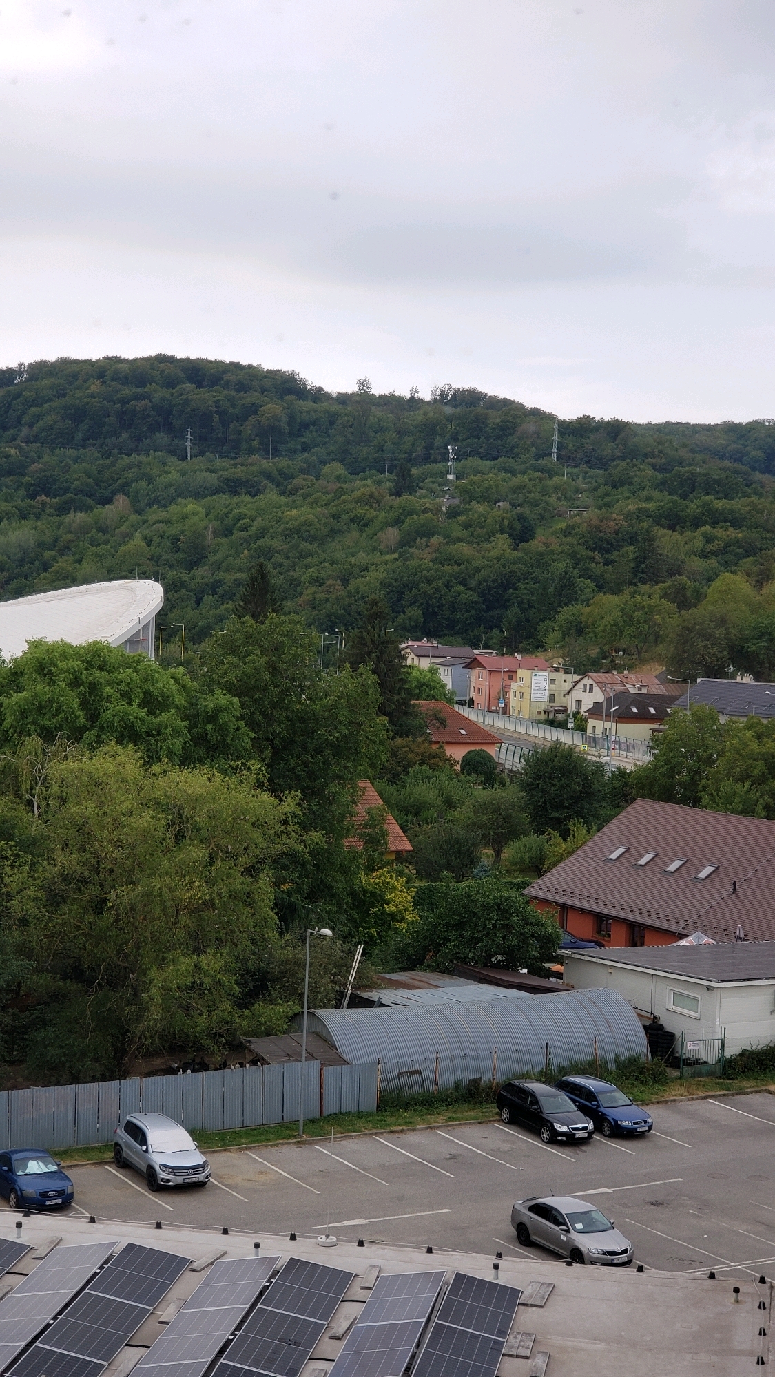 The image shows a view of a residential area and a wooded hill in Prešov, Slovakia. In the foreground, a rooftop is covered with solar panels. Below the rooftop, there is a parking lot with several cars, including a blue sedan, a silver SUV, a black hatchback, and a silver sedan. Behind the parking lot, a corrugated metal structure is visible, as well as a grey metal fence. Further in the background, a cluster of houses with red and beige roofs are situated amongst trees. A white building with a curved roof, possibly a stadium or sports complex, is located to the left of the houses. The distant hill is densely covered with green trees, and several telecommunication towers are visible on its slope. The sky is overcast.