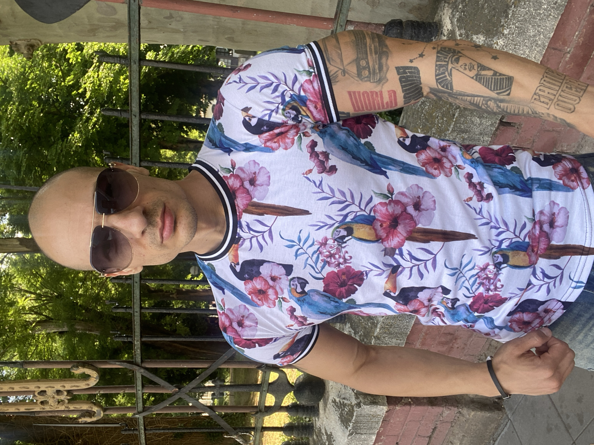A bald man with stubble and aviator sunglasses stands outdoors, looking directly at the camera in Pleven, Bulgaria. He wears a white t-shirt featuring a vibrant tropical print of blue macaws, black and white toucans, and pink hibiscus flowers, with black and white striped trim on the collar and sleeves. His left arm is extensively tattooed with images including a classic car, an Egyptian pharaoh's head, and partial text like "GAME OVER" and "WORLD". A black bracelet adorns his right wrist. He is positioned in front of a dark, ornate metal fence, behind which lush green trees are visible under bright sunlight. A low stone and brick wall runs along the bottom of the scene.