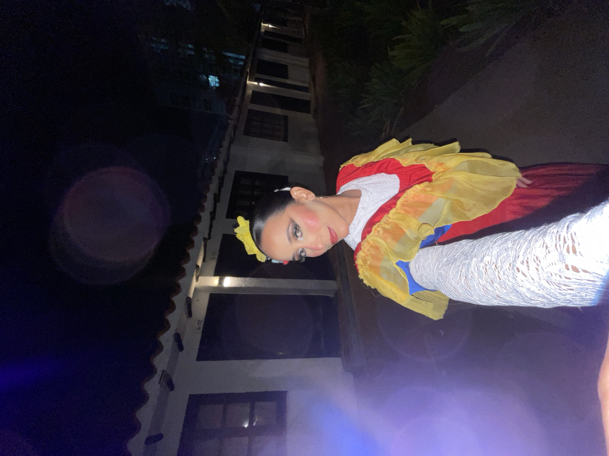 A woman dressed in a traditional Colombian folk costume is standing outside at night in front of a row of stucco buildings. The costume includes a red dress, a yellow ruffled overlay, and a white lace sleeve. The woman has her hair pulled back and is wearing makeup. The buildings appear to be motel rooms, with doors labeled "G-12" and "G-13". There are plants in the foreground and background, and the scene is lit by artificial lights. The woman is holding a camera and appears to be taking a selfie. The overall scene suggests a cultural event or performance taking place in Porlamar, Venezuela.