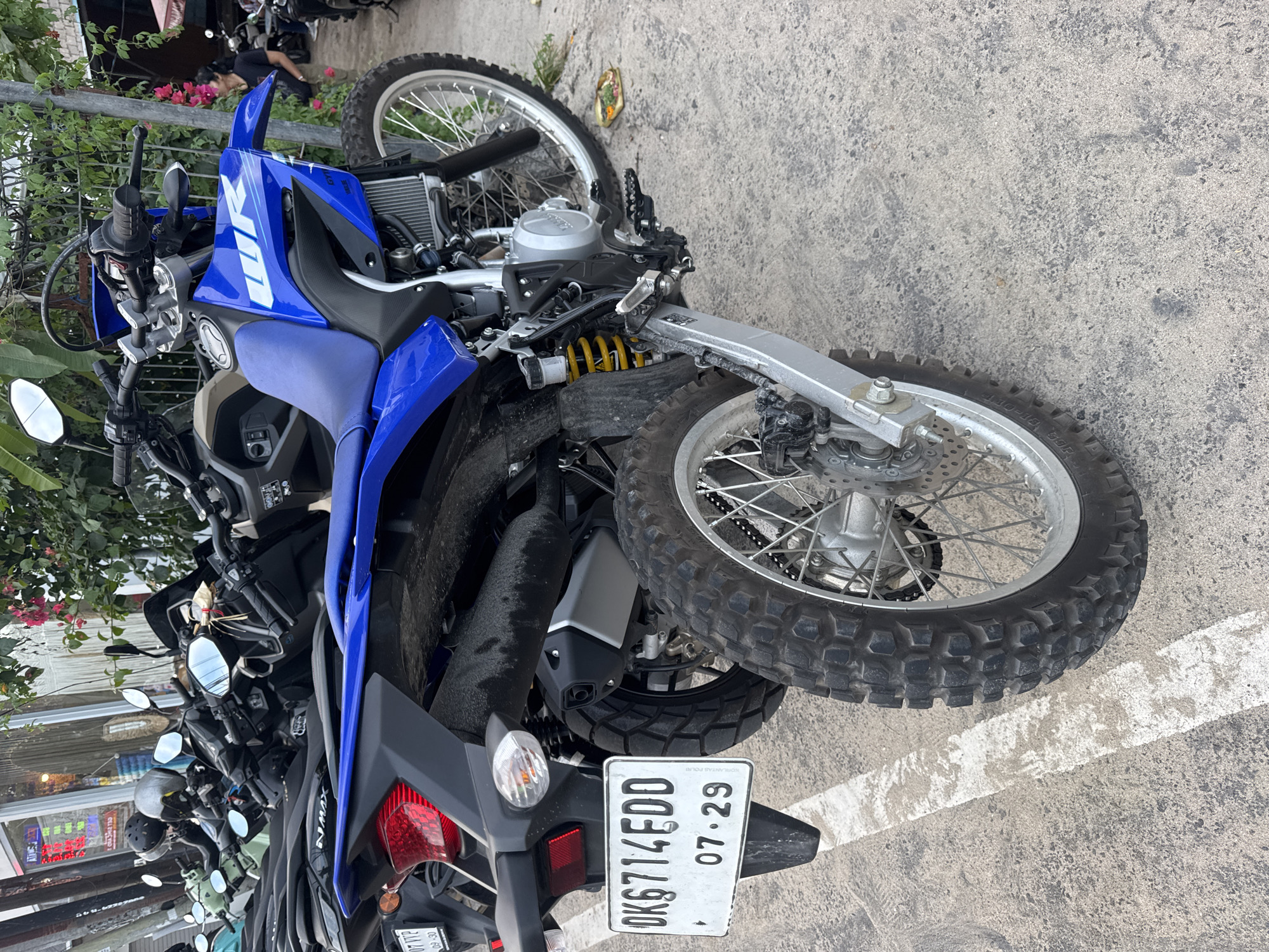 In Kuta Selatan, Indonesia, a blue Yamaha WR155R motorcycle is parked on a concrete surface marked with a white stripe. The motorcycle is positioned facing away from the viewer, with its rear tire prominent in the foreground. The license plate, "DK6714EDD 07.29," is clearly visible on the rear fender. Other motorcycles are parked behind it, with some of their details visible, suggesting a parking area. A woman wearing a black t-shirt and tan pants is partially visible in the background, standing near a cluster of pink flowers and foliage. The overall scene depicts a street-side or parking area, common in the bustling environment of Kuta Selatan.
