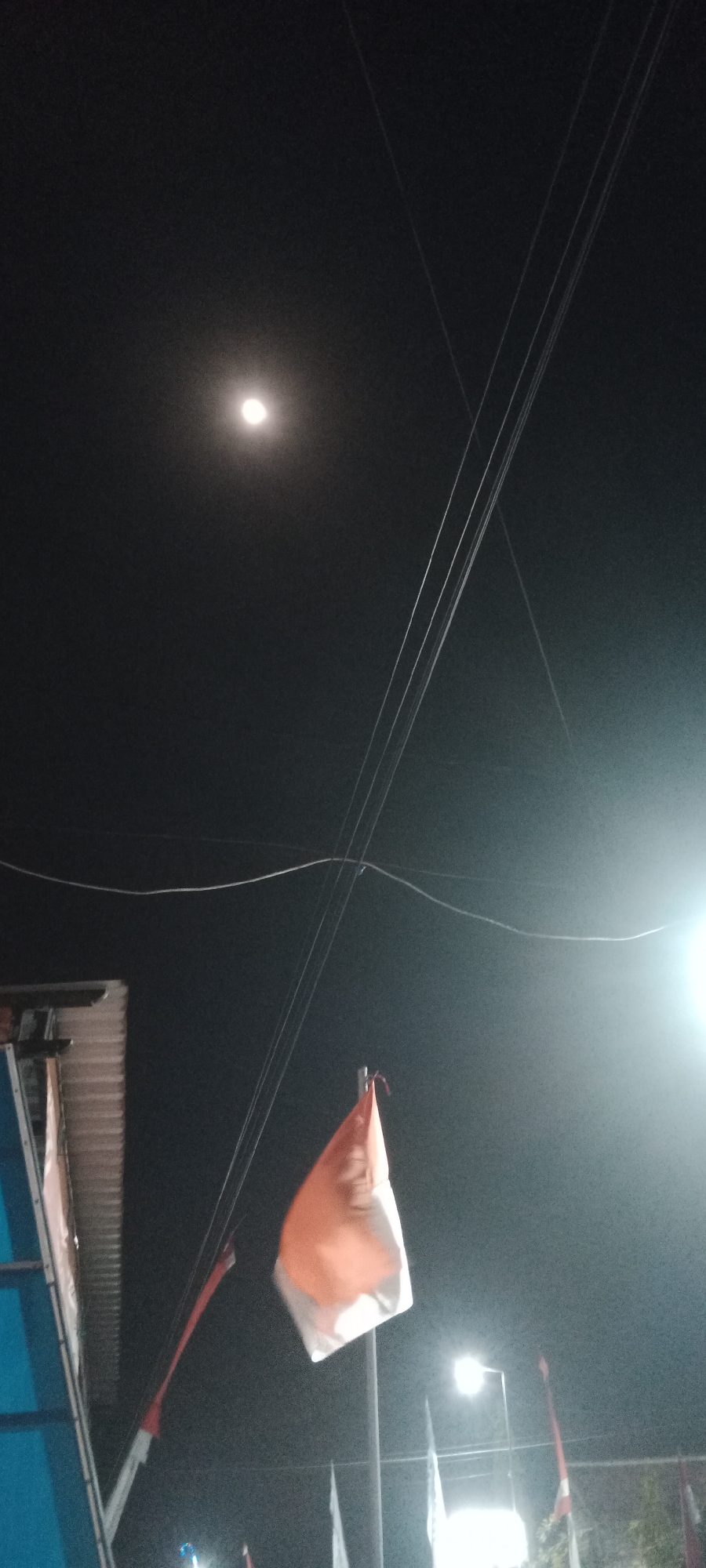 This is a low-light, vertical, outdoor photograph taken at night. The sky is dark, with a bright, full moon visible in the upper left quadrant. Several power lines are strung across the frame, appearing as thin white lines against the dark sky. In the lower center of the image, an orange and white flag is flapping in the wind, attached to a pole. To the left of the flag, a corrugated metal roof structure is visible. In the lower right, a bright street lamp illuminates the scene, with other flags and poles also faintly visible in the background. The overall impression is one of a festive or outdoor event taking place at night in an urban or semi-urban environment. No specific landmarks indicating the location of Pelang, Indonesia, are discernible in this image.