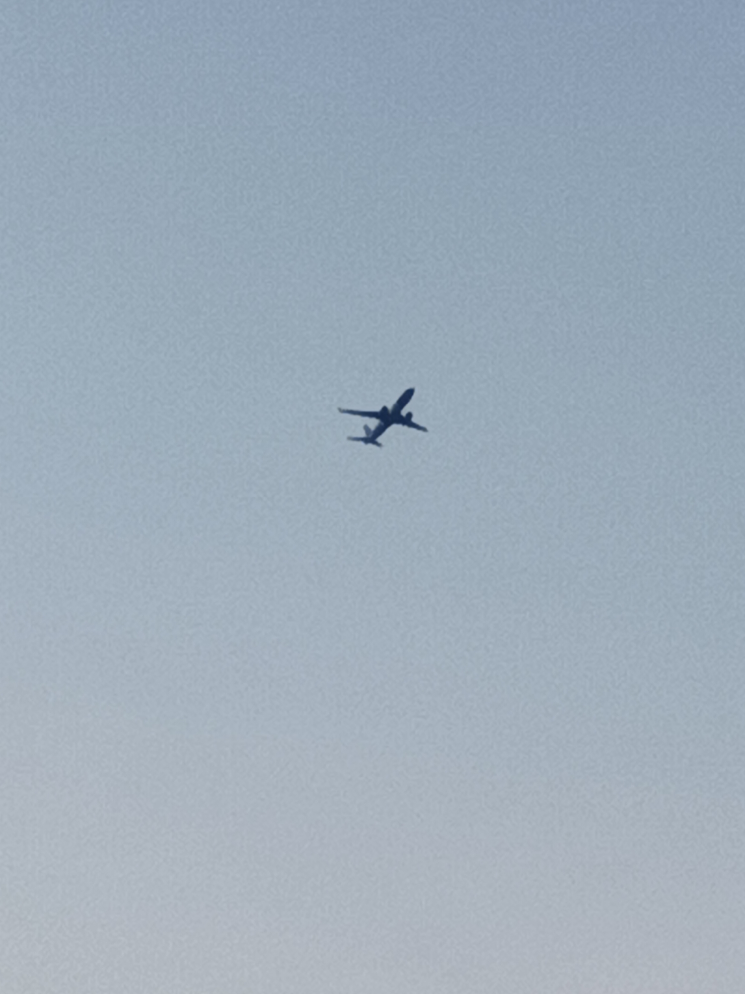 An airplane is seen flying in a clear, light blue sky. The airplane is a dark silhouette, appearing to be a commercial jet with two engines visible under its wings. It is angled slightly upwards, indicating it is climbing or in level flight. The image is a straightforward depiction of an aircraft in the sky, with no discernible background elements that would indicate a specific location within Prague, Czechia.