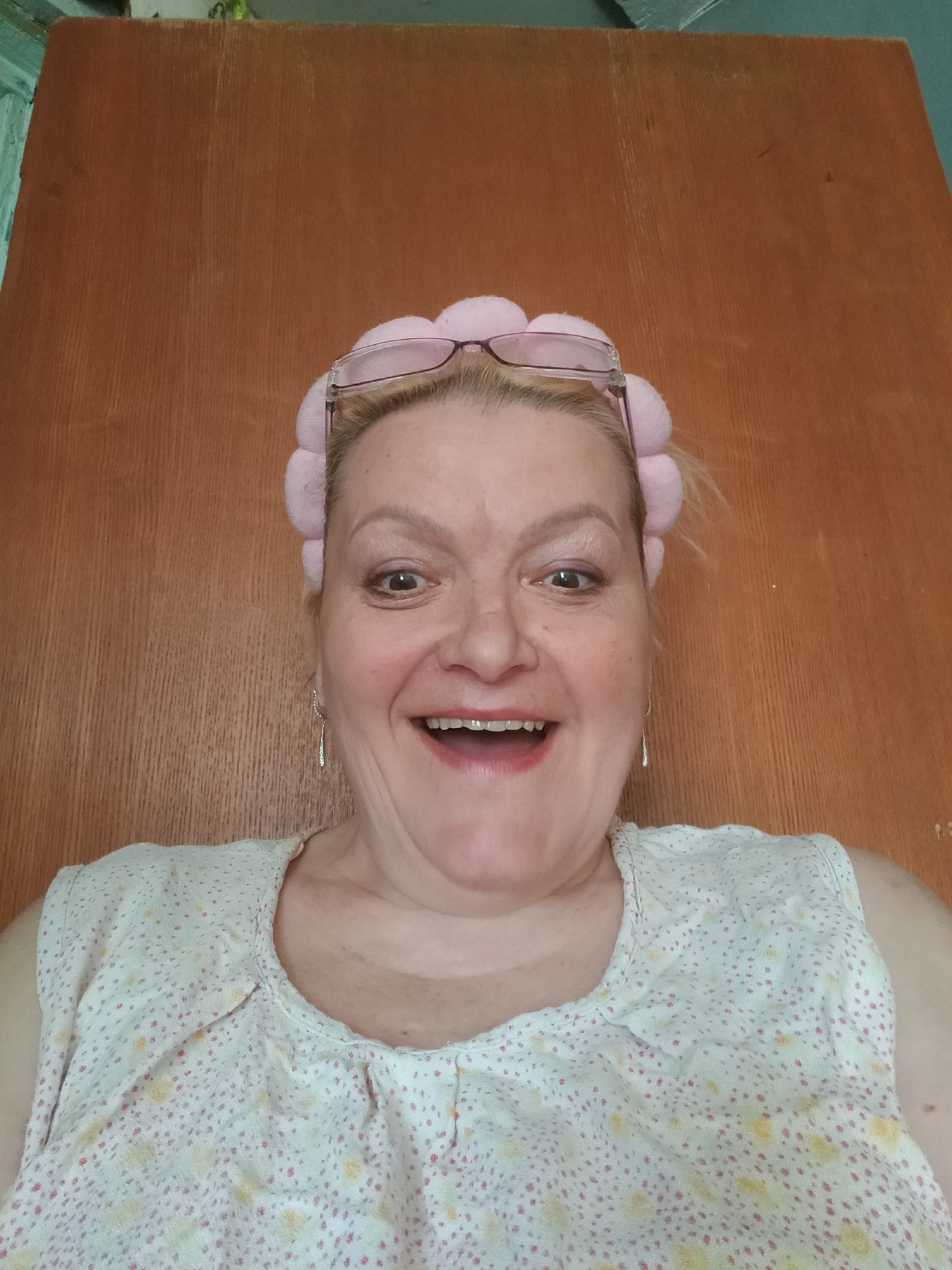 A medium close-up shot shows a middle-aged Caucasian woman smiling directly at the camera. She is wearing a pink headband with a row of rounded puffs and eyeglasses on top of her head. She has light blonde hair and fair skin with some freckles and wrinkles. Her teeth have metal braces on them. She is wearing a light-colored sleeveless top with a small floral print in pink and yellow. The background is a plain wooden door. There is no contextual information to place the scene in Красный Маяк, Russia.