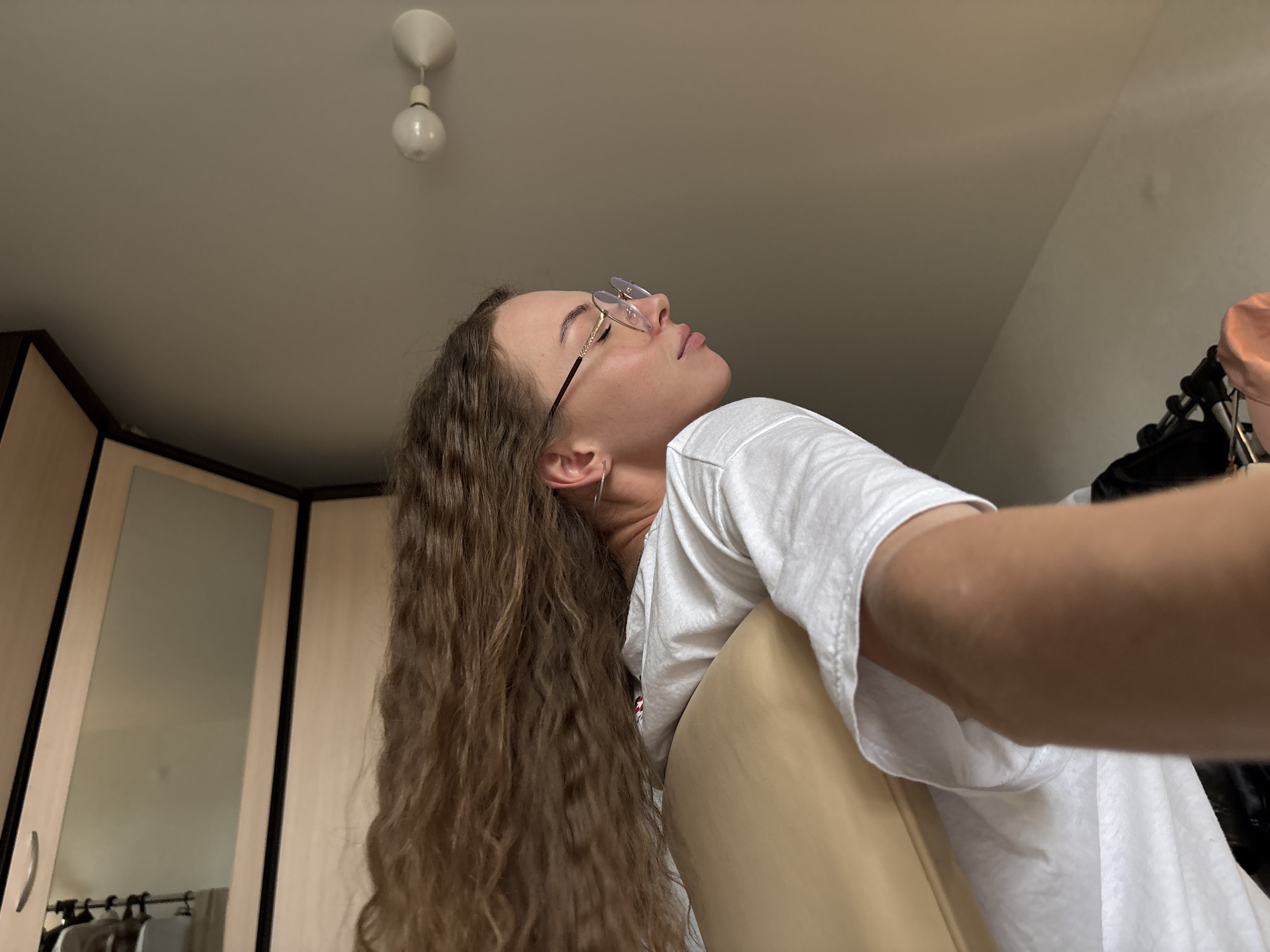 The image captures an indoor scene, possibly a bedroom, with a young woman in the foreground. She is wearing a white t-shirt and glasses, with her head tilted back and eyes closed, seemingly in a relaxed or contemplative pose. Her long, curly brown hair cascades down her shoulder. To the left of the frame, a portion of a wardrobe with mirrored doors is visible, reflecting some clothing items on hangers. A ceiling light fixture is also present in the upper portion of the image. The overall lighting suggests an indoor environment, possibly during the daytime, with natural light filtering in. The location in the city of Perm, Russia, is not discernible from the visual information provided in the image.