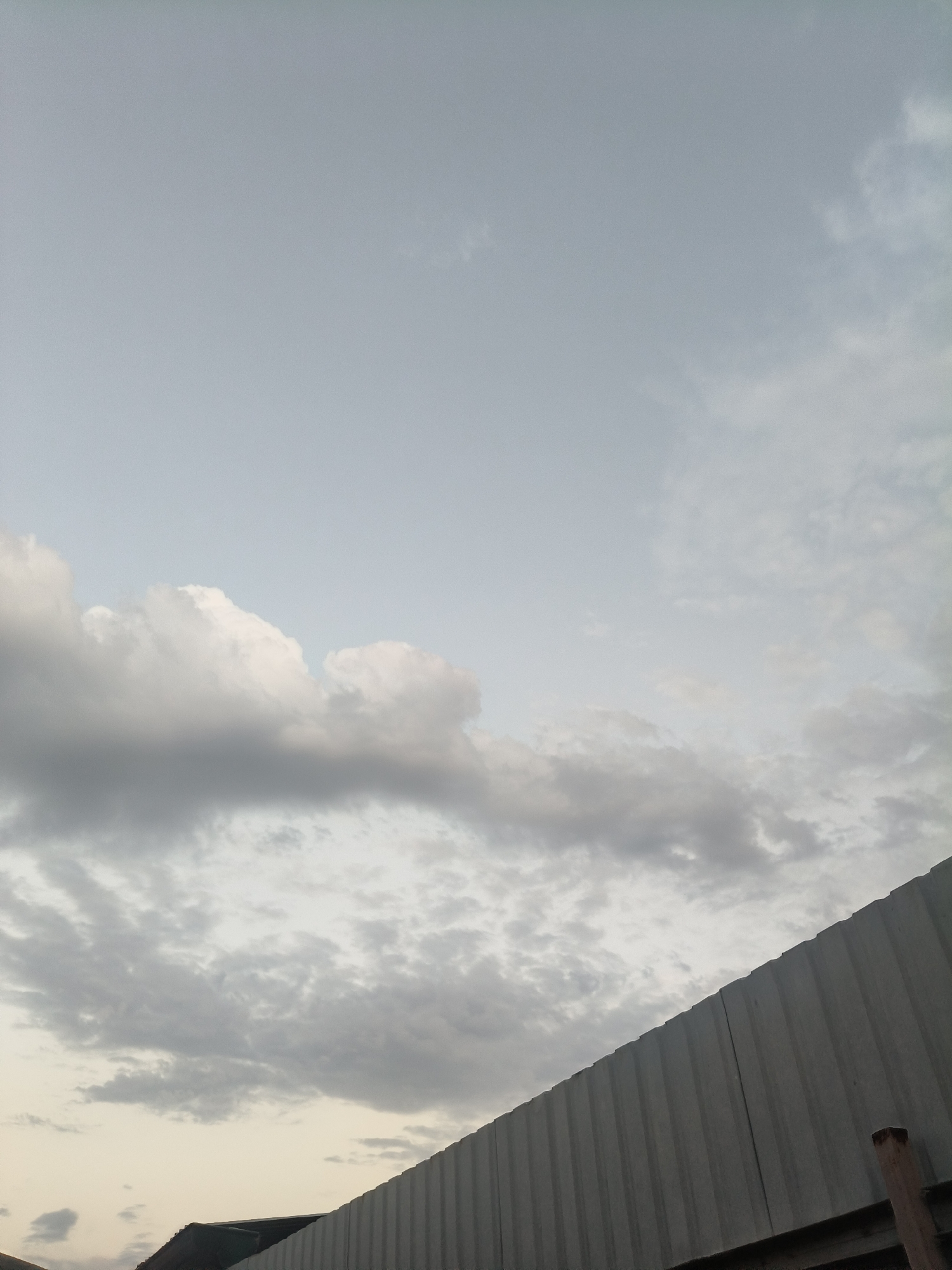 The image shows a cloudy sky with a corrugated metal structure in the foreground. The sky is a pale blue with various shades of gray clouds, some appearing fluffy and white, others darker and more diffuse. The metal structure runs diagonally across the bottom right of the frame, appearing to be a fence or a wall. A portion of a dark roof is visible beneath the metal structure on the left side. The overall impression is that of an overcast day, possibly near sunset or sunrise given the soft light. The location is listed as Unknown, Unknown.