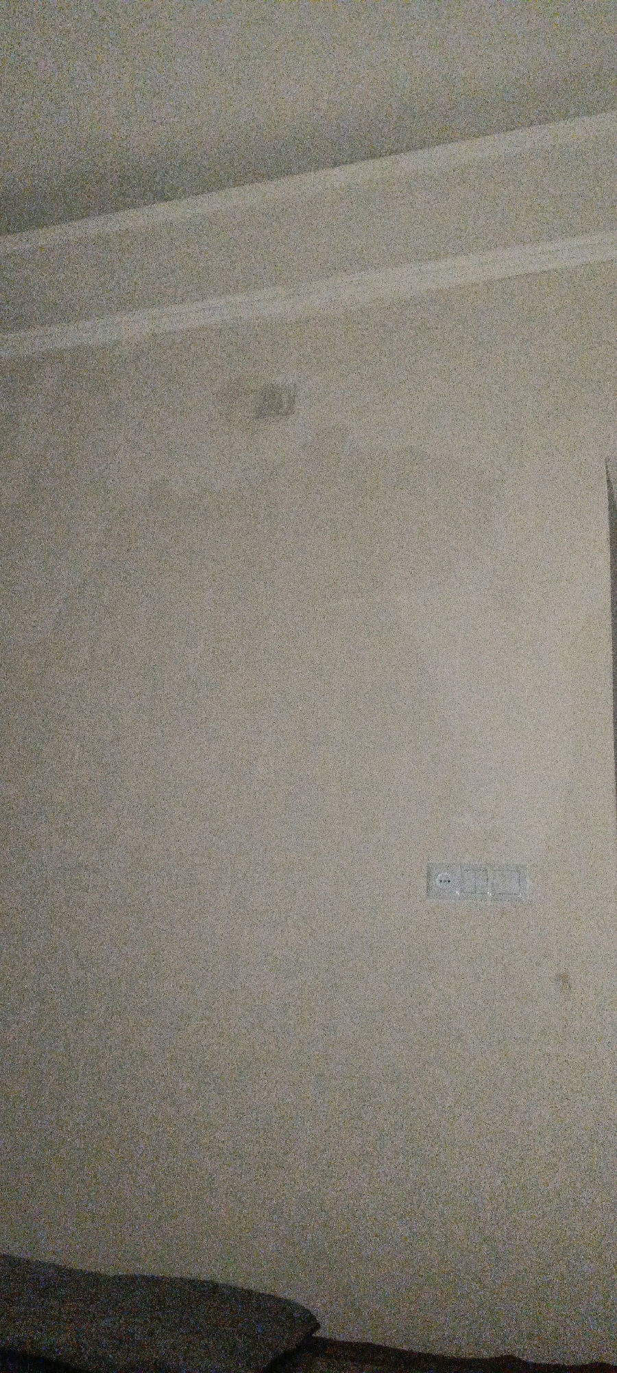 The image shows a lightly textured wall in a room, likely a domestic setting. The wall color is a neutral tone, possibly a pale beige or off-white. In the upper left quadrant, there is a subtle, slightly darker patch on the wall, suggesting a possible stain or discoloration. Towards the right side of the image, at mid-height, a standard electrical outlet with three modules is visible. The bottom of the image shows a dark, textured surface, possibly furniture or a carpet. The lighting appears to be ambient, casting a soft, diffused light across the scene. The image is grainy, suggesting it was taken in low light conditions or with a low-resolution camera. The location is identified as Рохинав, Tajikistan.