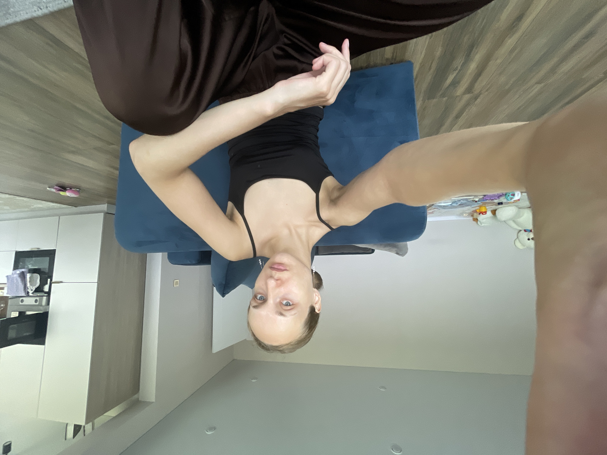 A young woman with blonde hair pulled back in a bun is sitting on the floor in a modern apartment, facing the camera. She is wearing a black tank top and dark brown wide-leg pants. Her left arm is extended towards the camera, as if taking a selfie. In the background, a large teal sofa is visible, along with a white kitchen cabinet unit with appliances and a stovetop. A white teddy bear and other toys are scattered on the floor near the sofa. The flooring is wood-look laminate, and the walls are painted a neutral light gray. The scene appears to be in a living area of an apartment in the city of Podshivalovo, Russia.