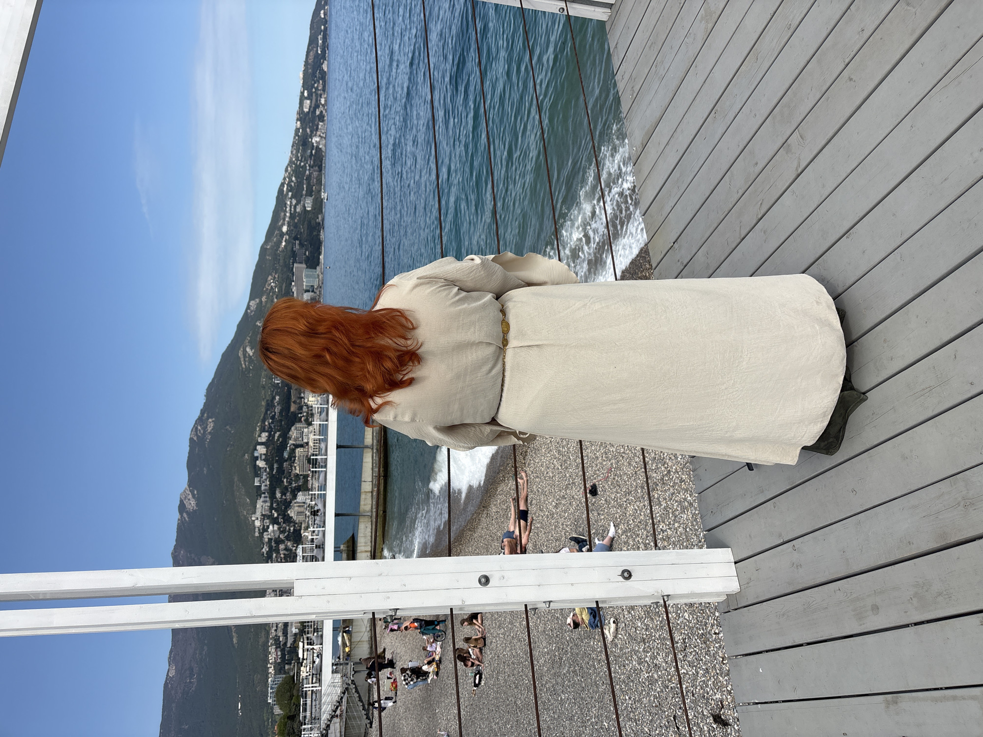A woman with red hair stands on a gray wooden boardwalk in Ялта, Ukraine, overlooking a pebble beach, the sea, and a mountain with buildings. She wears a long, light-colored dress and faces away from the camera. The sea is blue with white waves near the shore. People are relaxing on the beach below. A white railing is visible on the left side of the frame. The sky is blue with some clouds.
