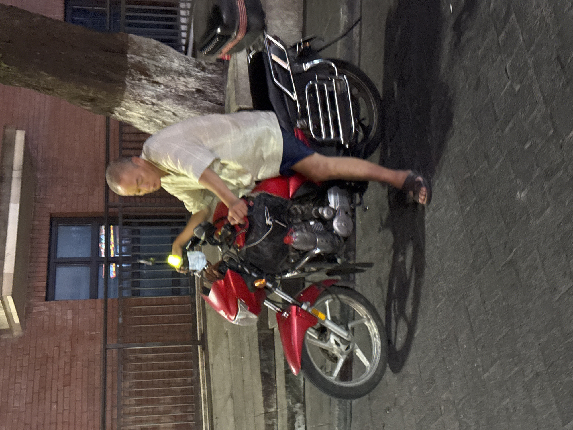In the Yuzhong District of China, an older Asian man is sitting on a red motorcycle. He is wearing a light-colored short-sleeved shirt and dark shorts, with dark sandals on his feet. A light-colored face mask is visible on the motorcycle's handlebars. The motorcycle has a black seat, a chrome luggage rack, and a black storage box on the back. The background consists of a brick building with windows and a metal fence, and a large tree with a white-banded trunk. The ground is paved with dark rectangular stones.