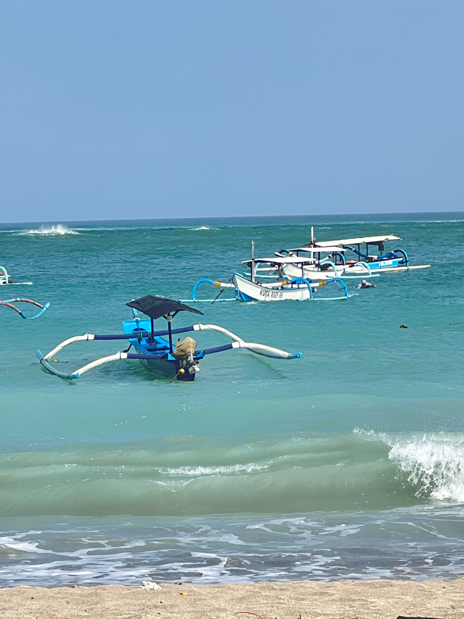 This image depicts a tranquil scene at Kuta Beach in Kuta, Indonesia, on a clear, sunny day. The turquoise waters of the Indian Ocean gently lap against the sandy shore, with a series of small waves breaking in the foreground. Several traditional outrigger boats, known as jukungs, are anchored in the shallow water. One blue jukung in the mid-ground is equipped with a black canopy and a tan bag or cooler. Further out, two larger white jukungs with blue trim are visible. One of these boats has "KUTA REEF 69" written on its hull, and a person is swimming in the water near it. The horizon is a clear blue sky, with no clouds visible. The overall impression is one of a peaceful day at a popular Indonesian beach destination.