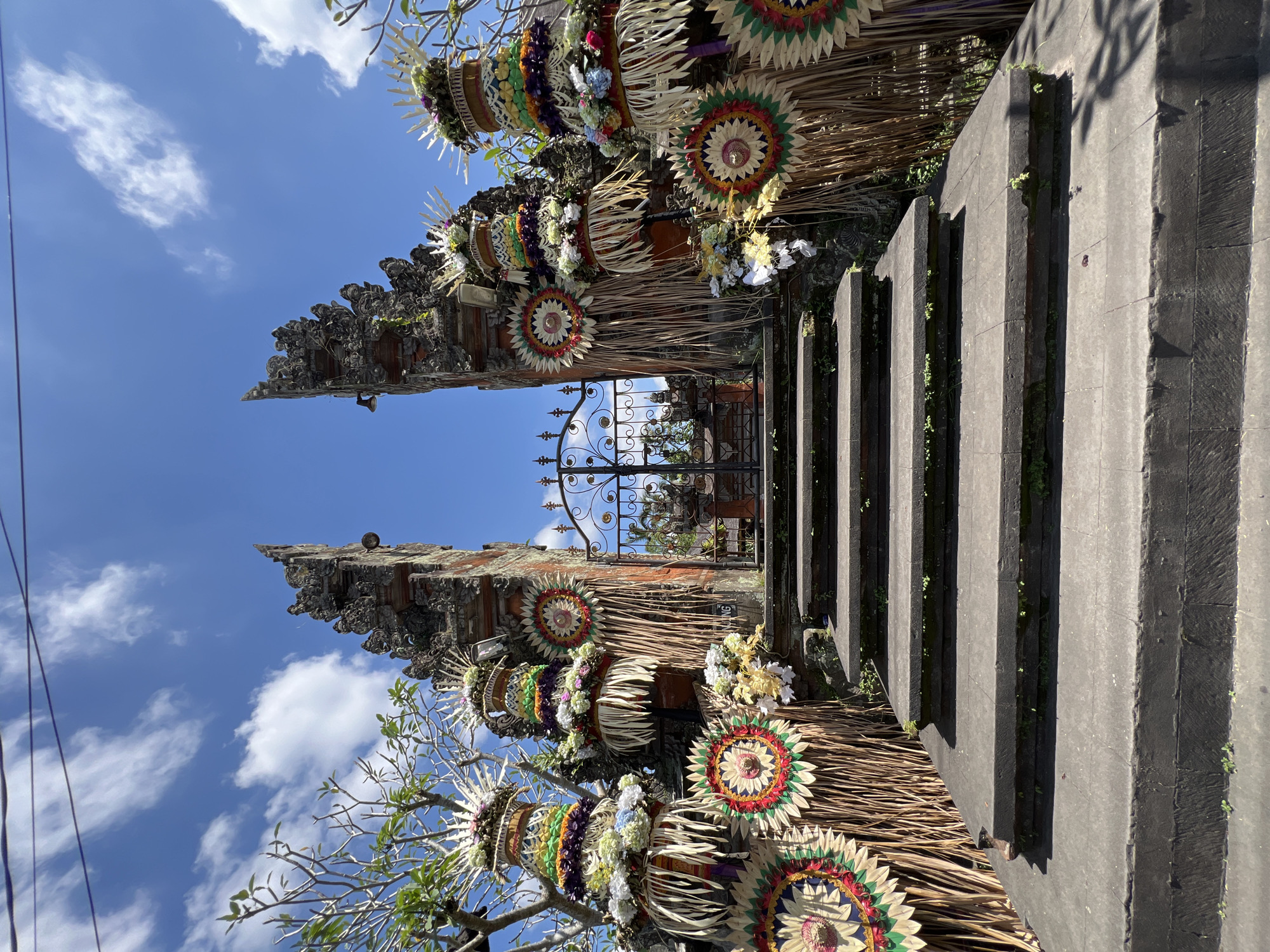 The image shows a traditional Balinese gate in Kedewatan, Indonesia. The gate is decorated with woven straw ornaments and colorful flowers. Stairs lead up to the gate, which is made of dark stone. The sky is blue with scattered white clouds. There is a wrought iron gate visible through the opening in the stone gate.