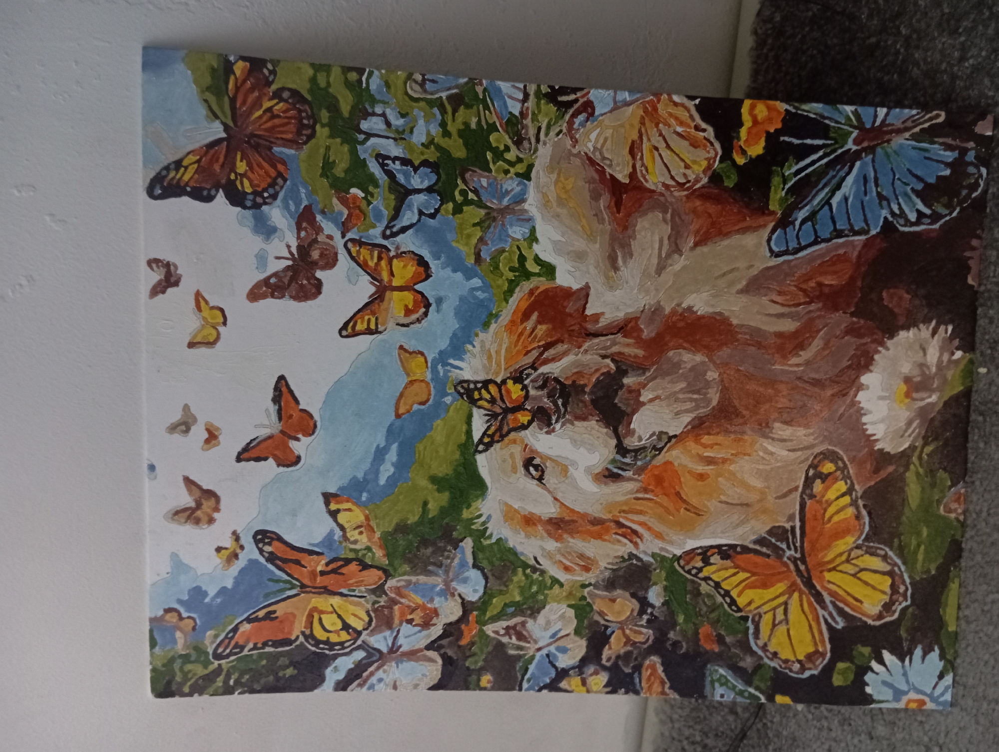 The image is a painting of a fluffy, light-colored dog with a butterfly perched on its nose. The dog is surrounded by numerous butterflies of various colors, including orange, yellow, and blue. The background depicts a landscape with green foliage and a light blue sky. The painting appears to be a "paint-by-numbers" artwork, with visible outlines and numbered sections within the paint strokes. The painting is set against a plain white wall and a textured gray surface, possibly a rug or couch. There is no discernible location within the city of Shamalgan, Kazakhstan.