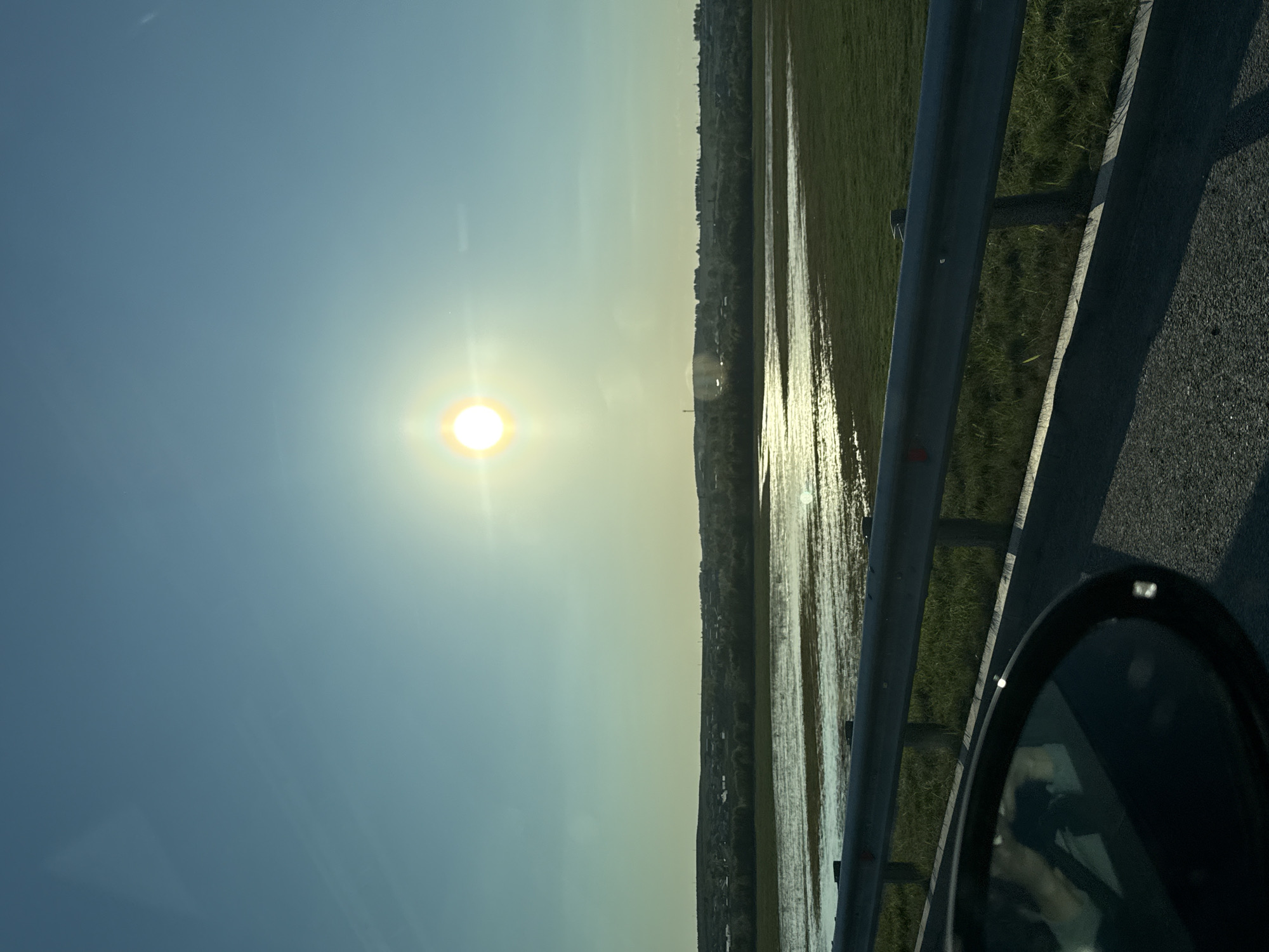 The image shows a view from a vehicle, possibly moving on a road. The sun is prominent in the sky, which transitions from light blue to a yellow-ish hue near the horizon. Alongside the road, there is a body of water reflecting the sunlight, potentially a flooded area or a lake, adjacent to some green land. A metal guardrail is visible next to the road. Part of the vehicle's side mirror is visible in the bottom right corner. The location is possibly in the area of Tarasovo, Russia, as indicated.