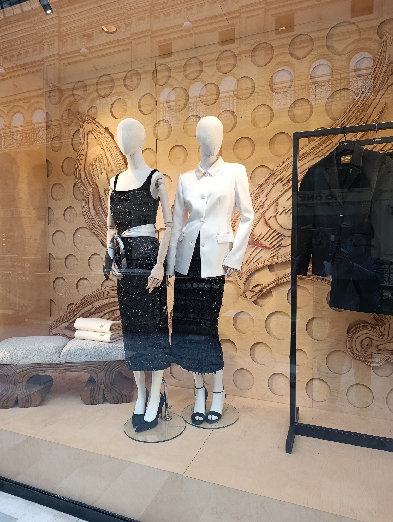 This is a full-shot image taken through a glass window, possibly a store display. The scene features two mannequins dressed in elegant attire. The mannequin on the left wears a sparkling black sleeveless dress with a white and black patterned sash tied around its waist. It is accessorized with black pointed-toe high heels. The mannequin on the right is dressed in a crisp white blazer with two front pockets, worn over a black skirt. This mannequin sports black heeled sandals.
Behind the mannequins, the backdrop consists of a wooden panel with a repeating pattern of large circles. Superimposed on this are decorative elements that resemble carved wood, with swirling, organic shapes. To the right of the mannequins, a rack holds a black jacket, and a person's reflection is visible in the glass, holding a phone and seemingly taking a picture. In the foreground, to the left, a portion of a grey upholstered bench is visible, with two folded white items, possibly towels or garments, placed on it. The overall environment appears to be indoors, likely a retail setting, with a light wooden floor and a reflective surface creating a sense of depth and visual interest.