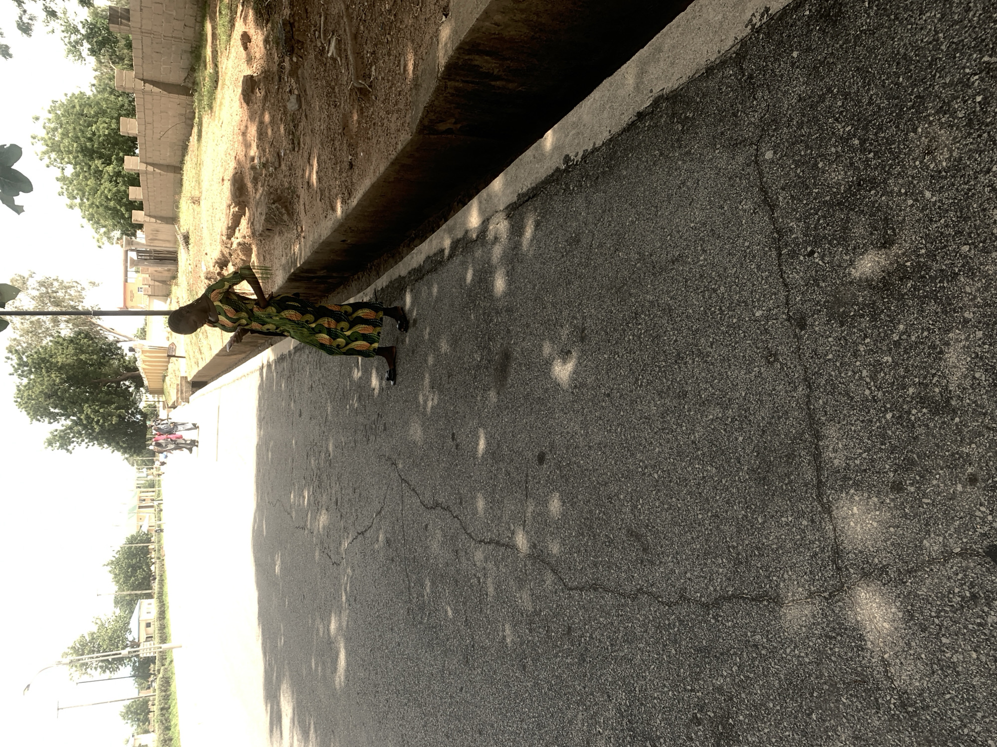 A person in a colorful, patterned dress stands on a cracked asphalt road in Jalingo, Nigeria. The road runs alongside a raised concrete planter box filled with dirt and sparse vegetation, beyond which is a low, sandy wall. Mature trees with green foliage line the area behind the wall, some casting dappled shadows on the road. In the distance, buildings and more trees are visible, suggesting an urban or semi-urban environment. The lighting indicates it is daytime, with shadows suggesting the sun is at an angle. The image is taken from a low angle, looking up towards the person. Several other people can be seen in the background, some on motorcycles. The overall scene depicts a typical streetscape in a warm climate.