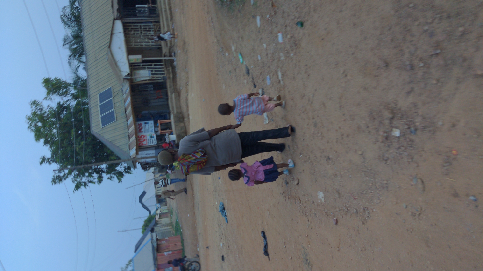 The image depicts an unpaved road in a residential area, likely on the outskirts of a city. A man, appearing to be an adult male, is walking along the road, holding hands with two young children, one on each side. The child on the right is wearing a pink dress and the child on the left is wearing a striped shirt. The man is wearing a light-colored shirt and dark pants. He is carrying a colorful bag slung over his shoulder.
To the left of the road, there is a building with a corrugated metal roof and a solar panel on its roof. There are signs displayed in front of the building, suggesting it is a business. Other structures and trees are visible in the background, as well as electrical lines overhead. The ground is covered in dirt and gravel, and there is scattered trash along the side of the road.
Although the image is not explicitly labeled as being in Masaka, Nigeria, the presence of a building with solar panels on the roof, the clothing of the subjects, the unpaved road, and the overall style of the architecture, the location is consistent with many scenes in Nigeria.