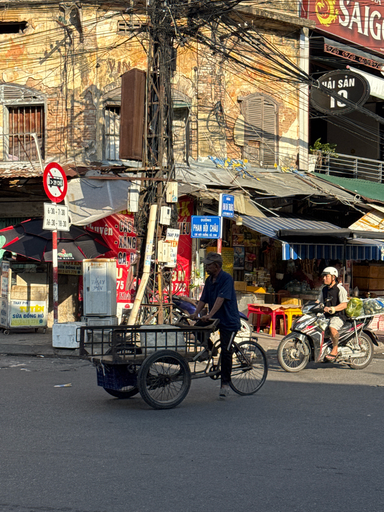 The image depicts a street scene in Nha Trang, Vietnam, characterized by its bustling urban environment. In the foreground, a man wearing a dark blue t-shirt and a baseball cap pedals a bicycle with a cargo trailer attached. The trailer is laden with a blue plastic bin and appears to be in motion, suggesting the man is engaged in transportation or delivery. Behind him, a motorbike with two riders passes by. The rider in front wears a white helmet and a black vest over a patterned shirt, while the passenger behind him is not clearly visible. The motorbike is carrying a large bag of green produce on the rear rack, indicating it is also involved in transport.
The backdrop of the scene reveals a dense network of electrical wires overhead, a common sight in Vietnamese cities, tangled around utility poles. The buildings lining the street show signs of age, with weathered facades and exposed brickwork, some featuring shuttered windows and balconies. Various signs are visible on the buildings, some in Vietnamese and others in English, indicating businesses such as a seafood restaurant ("HẢI SẢN") and an auto repair shop. Traffic signs, including a red and white circular sign prohibiting buses and a rectangular sign with Vietnamese text and time slots, are also present, regulating the flow of traffic. The overall atmosphere suggests a busy commercial street with active local commerce and daily life unfolding. The lighting indicates it is daytime, with sunlight casting shadows on the street and buildings.