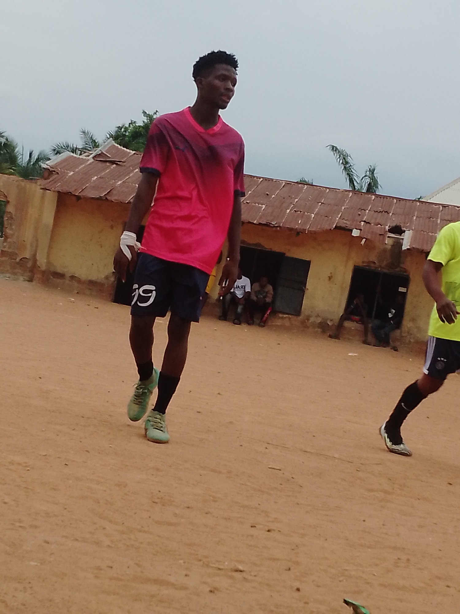 The photo depicts a young man walking on a dirt field in what appears to be Akwanga, Nigeria, based on architectural and environmental cues. He is wearing a pink and black t-shirt, dark shorts with the number "19" on them, and green cleats. His left hand is bandaged. Behind him, a dilapidated, single-story structure with a rusty, corrugated metal roof is visible. Several people are sitting or standing near the building's entrances, including another man wearing a yellow t-shirt. The sky is overcast.