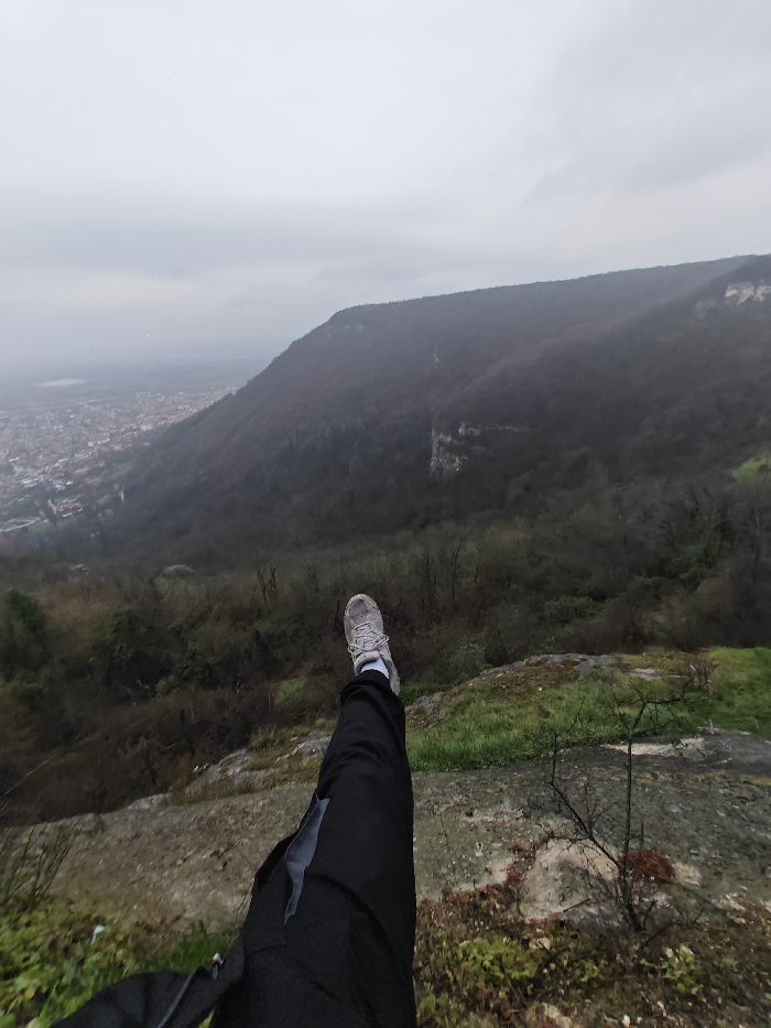 The image displays a panoramic view from a high vantage point on the Shumen Plateau, overlooking the city of Shumen, Bulgaria. In the lower-left foreground, the extended left leg and foot of an individual are visible, positioned over a steep drop-off. The leg is clad in dark-colored trousers featuring a lighter grey panel on the thigh. The foot wears a light grey athletic shoe with white laces and a visible light-colored sock. The immediate ground consists of rocky terrain partially covered by green moss and sparse brown vegetation. Below, a deep valley is densely forested with leafless deciduous trees. In the background to the left, a portion of the city of Shumen is visible, showing numerous buildings with varying roof colors, partially obscured by atmospheric haze. A distant, forested mountain ridge, also primarily composed of leafless trees and some exposed rock formations, forms the horizon. The sky is uniformly overcast and grey.