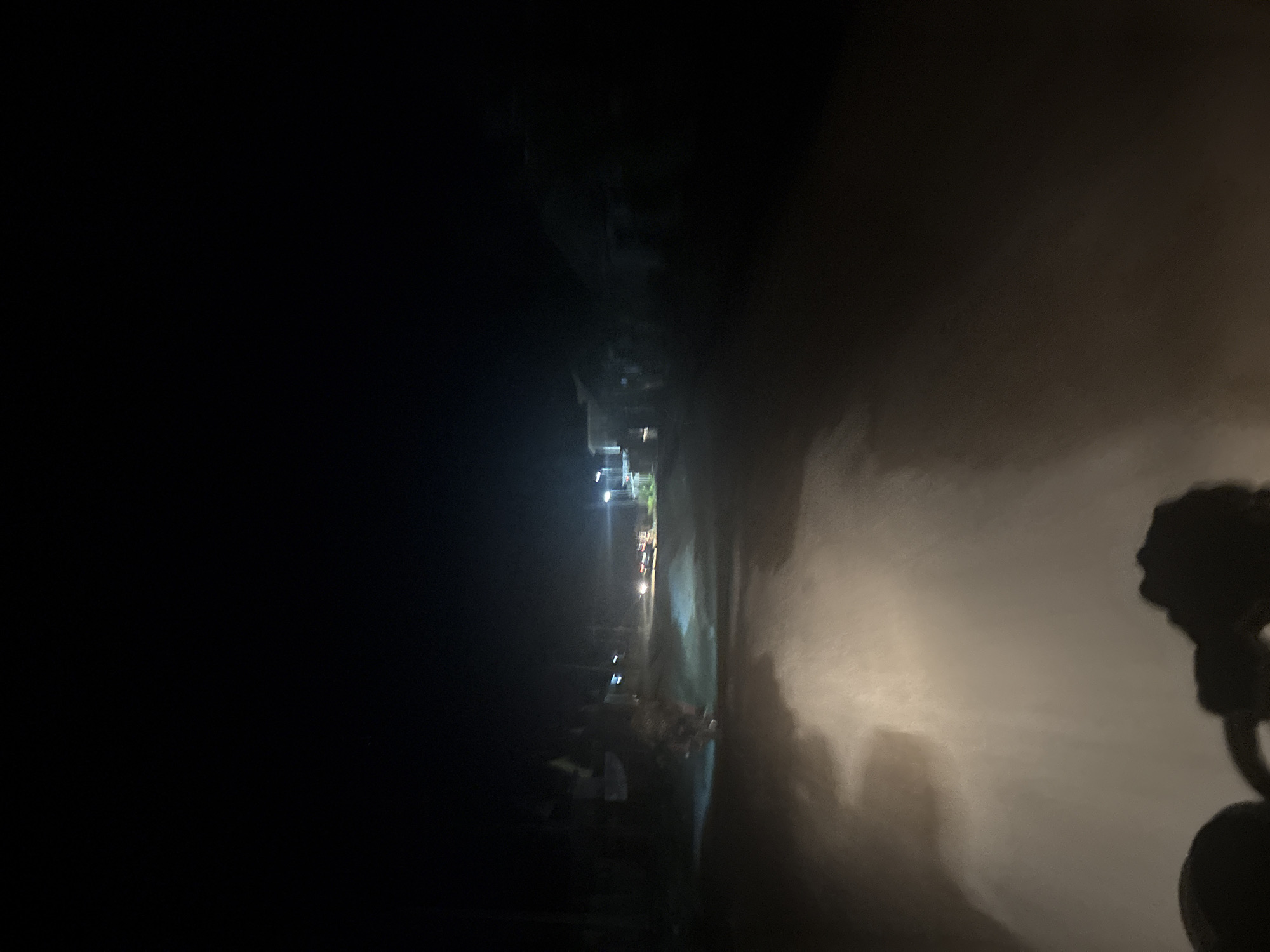 A nighttime scene captured from the vantage point of a moving vehicle, likely a motorcycle, traveling on an unpaved road in Angwan Sayawa, Nigeria. The lower portion of the image displays the dark silhouette of vehicle handlebars in the bottom-center and left. A powerful, warm-toned light source, consistent with a headlight, illuminates the immediate road surface, revealing light brown, dusty ground with visible patterns of disturbed particulate matter. The unpaved road extends into the midground, flanked by dimly visible, dark structures on both sides, presumably buildings. Multiple artificial light sources, including distant streetlights, are present further down the road, casting diffuse illumination. Two pairs of headlights from other vehicles are faintly visible in the distance. A noticeable atmospheric haze or dust diffuses the distant light sources. The upper two-thirds of the image consist of an undifferentiated, deep black expanse, indicating a dark night sky. No individuals are discernible.