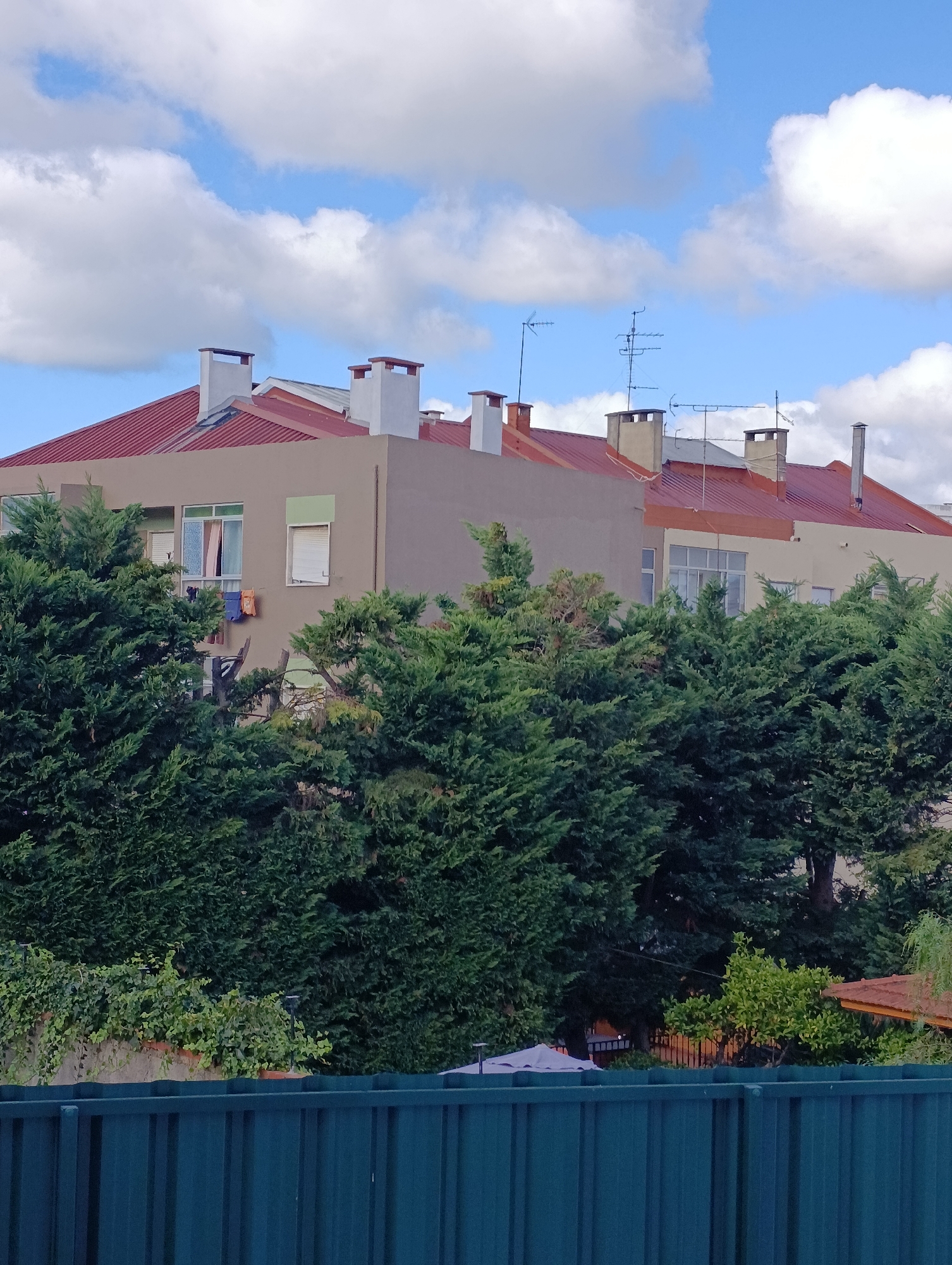The image shows a multi-story building with a red tiled roof, numerous chimneys, and several television antennas on its upper sections. The building's facade is a muted beige color. Visible on the exterior are windows, some with blinds, and clothes hanging on a line outside one window. Dense evergreen trees partially obscure the lower part of the building. In the foreground, a dark teal metal fence runs horizontally across the bottom of the frame. The sky above is blue with scattered white clouds. The scene appears to be a residential or apartment building.