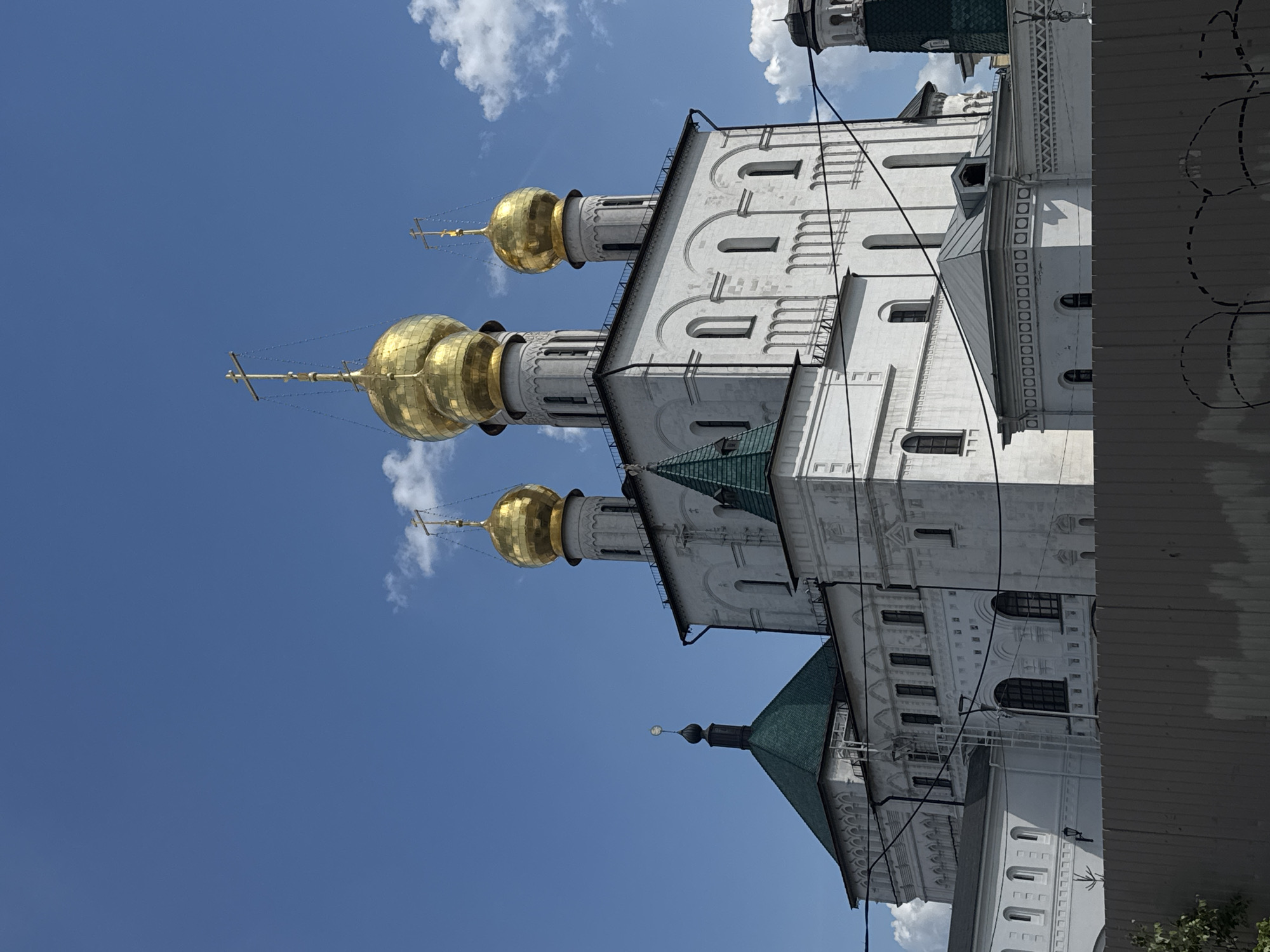 The image captures a majestic architectural complex, likely a Russian Orthodox church or cathedral, under a clear blue sky with scattered white clouds. The prominent feature is a tall, white building adorned with multiple gleaming golden onion domes, each topped with a cross.
The main structure showcases white stone or stucco walls, featuring several arched windows and intricate decorative moldings along its cornices and facades. At least three large golden domes are visible, rising above slender white towers that also have arched window openings. Parts of the roof are dark, possibly green, contrasting with the white walls and golden domes.
The bright sunlight illuminates the scene, making the golden domes reflect brightly. Several dark lines, likely utility wires or cables, are visible against the sky and partially draped across the building's facade. To the right of the main building, a darker, possibly corrugated metal, structure or wall is partially visible, featuring some faint, scribbled white markings.
The photograph is taken from a low angle, looking upwards at the imposing architecture, emphasizing its height and grandeur. The weather appears sunny, indicating daytime. There are no people visible, and no specific event is taking place beyond the static view of the edifice. The architectural style, particularly the onion domes, is characteristic of Eastern Orthodox churches. No visible text is present in the image.