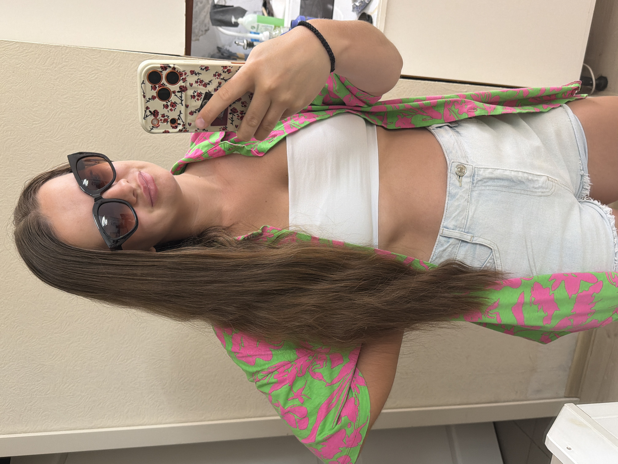 A young woman with long, dark brown hair is taking a selfie in a room. She is wearing large, black cat-eye sunglasses, a white strapless top, a bright green shirt with a pink floral pattern, and light blue denim shorts. She is holding up a smartphone with a floral case to take the picture. Her right arm is bent at the elbow, and her left hand is on her hip. She has a black braided bracelet on her left wrist. The background appears to be an indoor setting, possibly a home or hotel room, with light-colored walls and cabinets visible. There are no specific Sochi landmarks or indicators to place the location within the city.