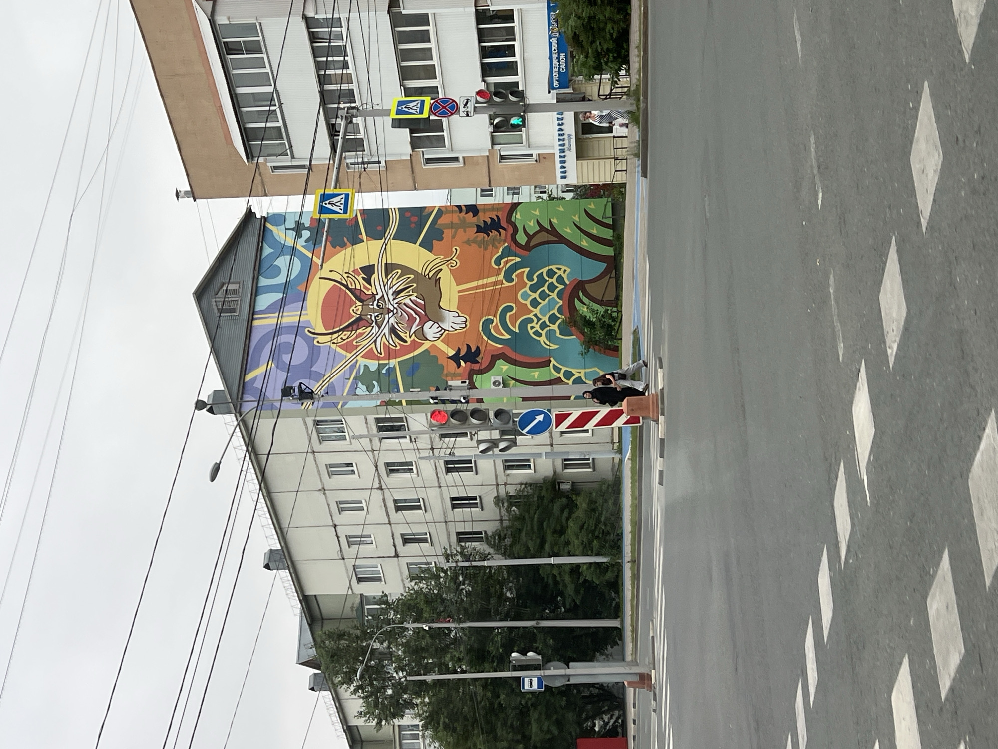 The image depicts a street scene in Yuzhno-Sakhalinsk, Russia, featuring a large mural on the side of a building. The mural, prominently displayed, is a colorful depiction of a stylized cat-like creature with large eyes and exaggerated whiskers, set against a background of a sun and swirling patterns.
In the foreground, a pedestrian crossing is visible on an asphalt road, with white markings indicating the crosswalk. Traffic lights are present, with one showing a red light and another showing a green light, suggesting an intersection. Several traffic signs are also visible, including a pedestrian crossing sign and a no stopping sign.
Adjacent to the mural, there are multi-story residential buildings. One building to the left appears to be a light-colored, low-rise apartment building. To the right of the mural is a taller, tan-colored building with balconies and white-framed windows.
Several power lines crisscross the sky above the scene. Trees are present on the left side of the image, providing some greenery. Two individuals are walking on the sidewalk near the traffic lights, and another person is walking near a storefront. The overall environment suggests an urban setting with a mix of residential and commercial elements. The sky is overcast, indicating a cloudy day.