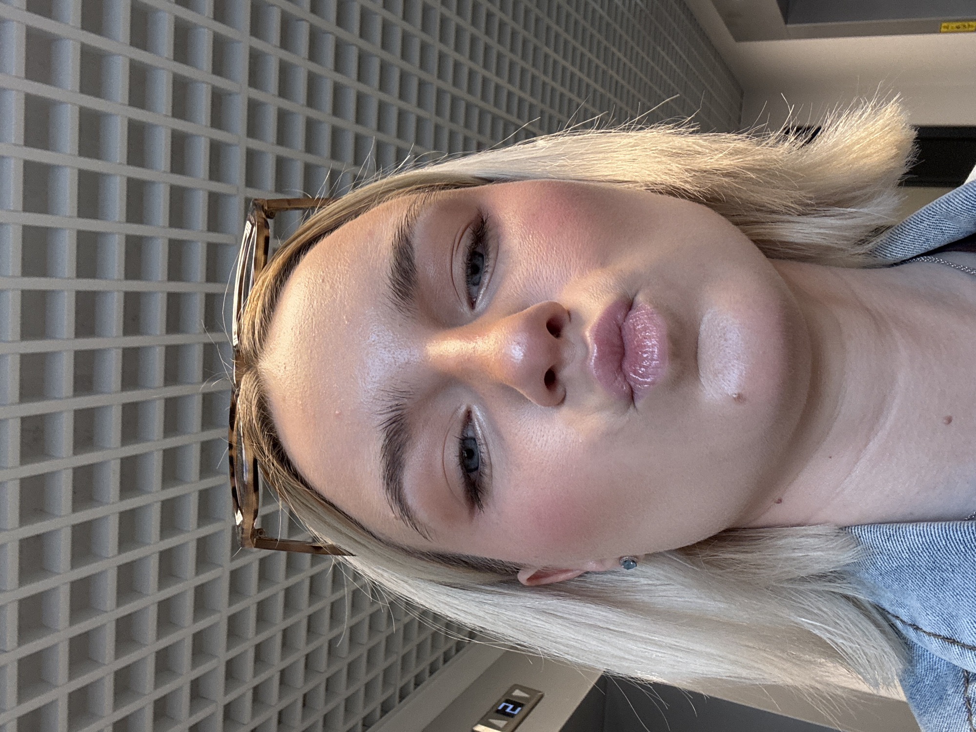 A close-up, angled portrait of a light-skinned person with blonde hair. The person is facing the camera with a slightly puckered expression. They have light blue eyes and visible pores on their skin. Glasses are perched on their head. The background features a grid-like pattern, possibly a building facade, and a glimpse of an elevator display showing the number "21". The person is wearing a denim jacket. Location: Unknown, Unknown.