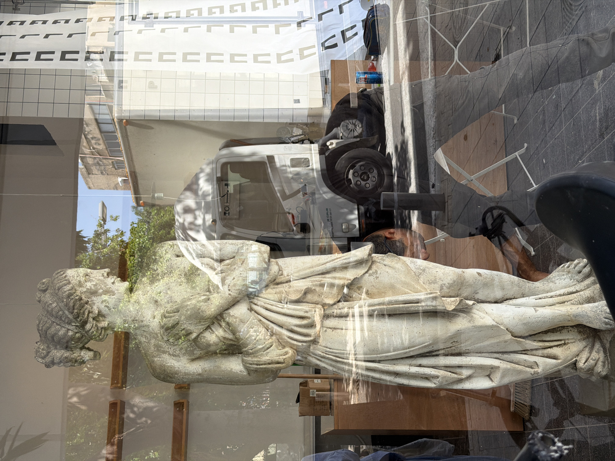The photograph depicts a statue of a classical figure inside a storefront in Tel Aviv, Israel. The statue is off-white and detailed, showing draped clothing and elaborate hair. It lies on a wooden surface inside the store, which is visible through a glass window.
The reflection in the glass reveals a scene outside the storefront. A white truck is visible, along with architectural details of buildings across the street. A man's head is also visible, presumably reflecting from the photographer's location. The background includes glimpses of trees and a tile exterior of a building.
