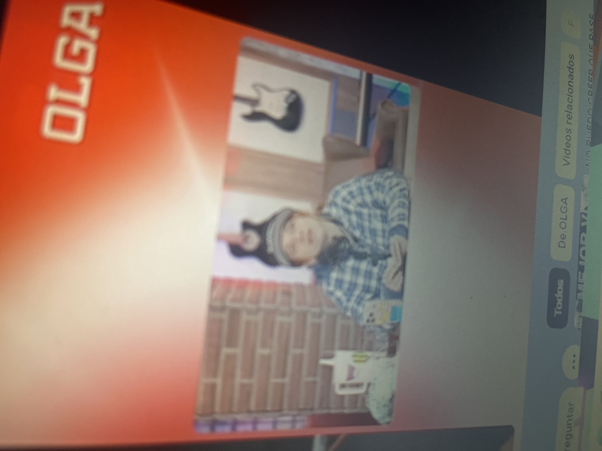 The image appears to be a screenshot or capture of a video stream, possibly from a computer or mobile device. At the top of the frame, a red gradient background features the word "OLGA" in large, white, capitalized letters. Below this, a smaller, pixelated video feed shows a person wearing a plaid shirt and a beanie, seated at a desk. To the left of the person is a white mug with a design. In the background, there is a brick wall and an electric guitar mounted on the wall. At the bottom of the frame, there are interface elements that resemble buttons or menu options, with text such as "Todos," "De Olga," and "Videos relacionados." The overall quality is low, with blurriness and visible pixelation. There is no identifiable information within the image that would place it in Santa Ana, Venezuela.