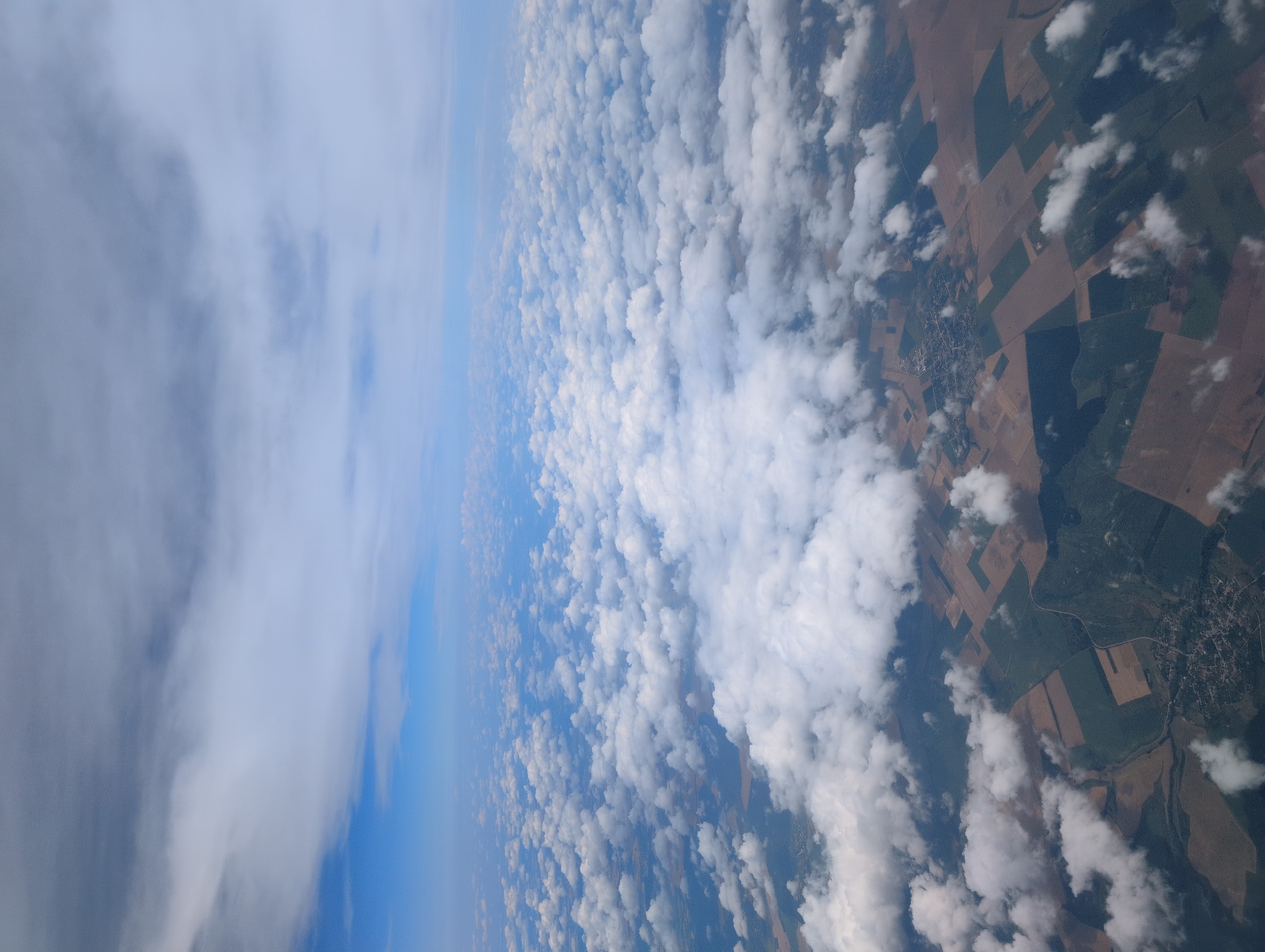 This is an aerial photograph, likely taken from an airplane, showing a landscape dotted with clouds. The view is from a high altitude, looking down at the earth and up towards the sky. The sky occupies the upper portion of the image and is a soft blue, with a variety of clouds. There are large, fluffy white cumulus clouds scattered across the scene, as well as thinner, more wispy cirrus clouds higher up, creating a layered effect.
The ground below is a mosaic of agricultural fields, varying in color from dark green to light brown and tan, suggesting different crops or stages of growth. Some areas appear to be forested, with denser patches of dark green. A small village or town is visible, with clustered buildings and what appears to be a road network. The fields are laid out in geometric shapes, typical of cultivated land. The interplay of sunlight and shadow from the clouds creates a dappled effect across the landscape, adding depth and texture to the scene. There are no people or discernible human activity visible in the image. The location is identified as the city of Luton, United Kingdom, although the specific features visible are common to many rural or semi-rural landscapes from this vantage point.