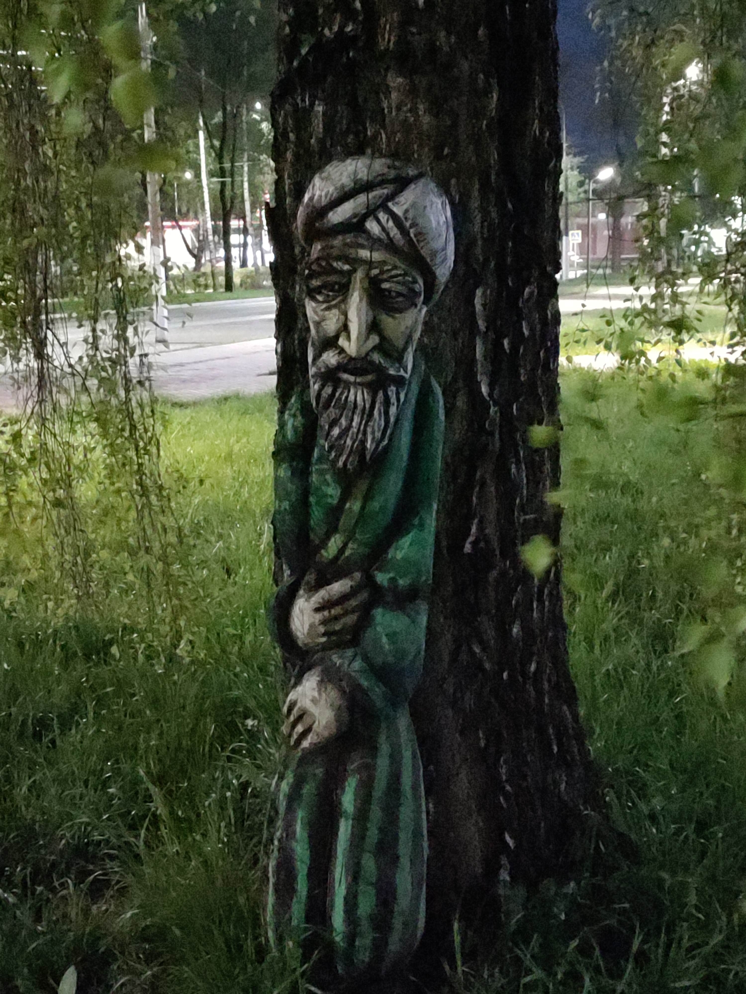 A carved wooden sculpture of a bearded man wearing a turban stands at the base of a tree. The sculpture is painted in green and gray, with dark vertical stripes on the lower portion. The tree is a dark-colored species, and the sculpture appears to be somewhat integrated into the tree trunk. Tall green grass surrounds the tree and sculpture. In the background, a road is visible, with street lights and buildings along the roadside. The location appears to be a park or grassy area near a street in a city, Unknown, Unknown.