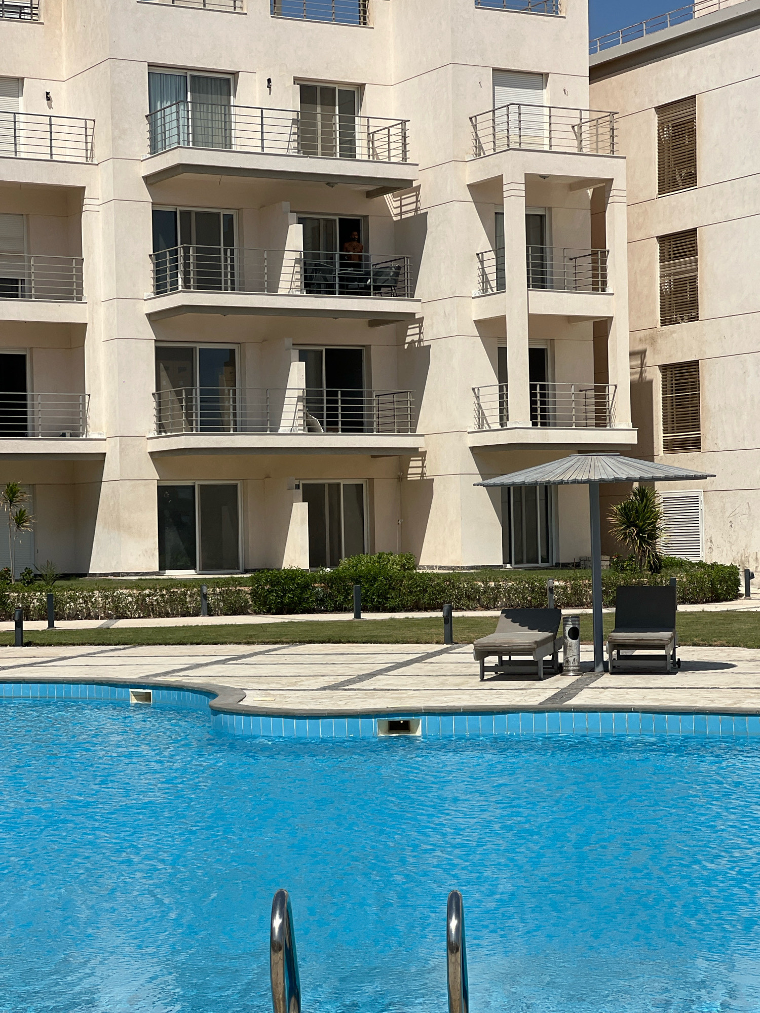 The image shows a modern apartment complex with balconies and windows, located in Sharm El Sheikh, Egypt. In the foreground, a bright blue swimming pool with metal railings is visible. The pool area is paved with light-colored tiles and features two lounge chairs and a large umbrella. Behind the pool, a manicured lawn and dense green bushes are arranged, with a few small outdoor lamps placed along the edge. The building's exterior is a light beige color, and the balconies are equipped with metal railings. One balcony on the second floor shows a man standing and looking out. The scene appears to be a resort or residential complex, likely in a sunny, warm climate.