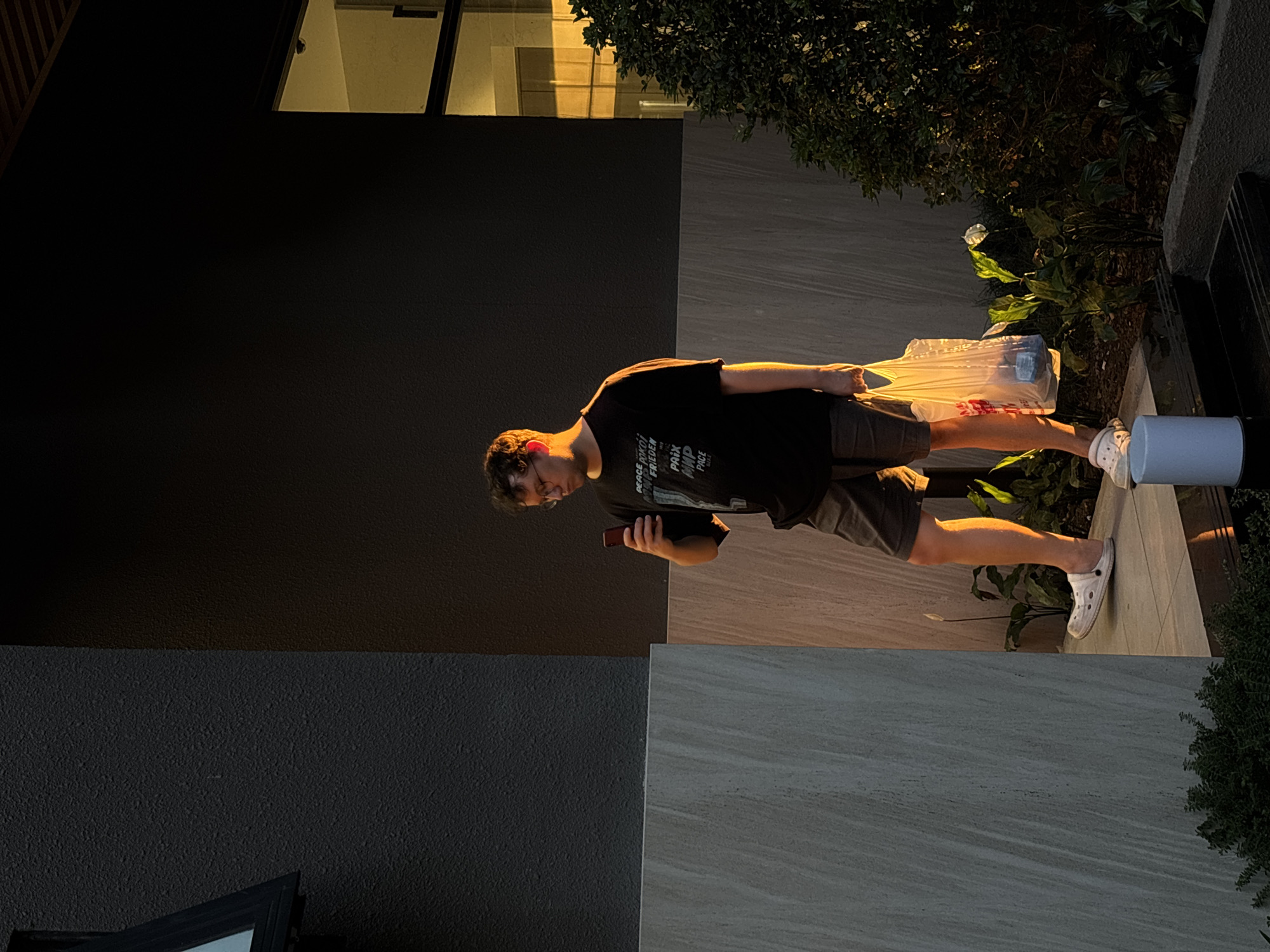 A young person is standing outdoors at dusk, holding a plastic bag filled with bottles. They are wearing a black t-shirt and dark shorts, along with white crocs. The t-shirt has text on it that appears to be in English and possibly French. The person is looking down at their phone, which they are holding in their right hand.
The scene is set against a building with a dark, textured wall and a section of lighter, stone-like tiles. Above, there's a glimpse of a window with light emanating from within, suggesting an indoor space. Lush green foliage borders the scene, indicating a natural element within the urban environment. The lighting suggests either early morning or late evening, with a warm glow illuminating the person's legs and the plastic bag, casting long shadows. The overall mood is casual and relaxed. The text on the t-shirt is partially visible and includes words like "PEACE," "TOKYO," "FRIEDEN," and "PRIX." The location context is Đà Nẵng, Vietnam.