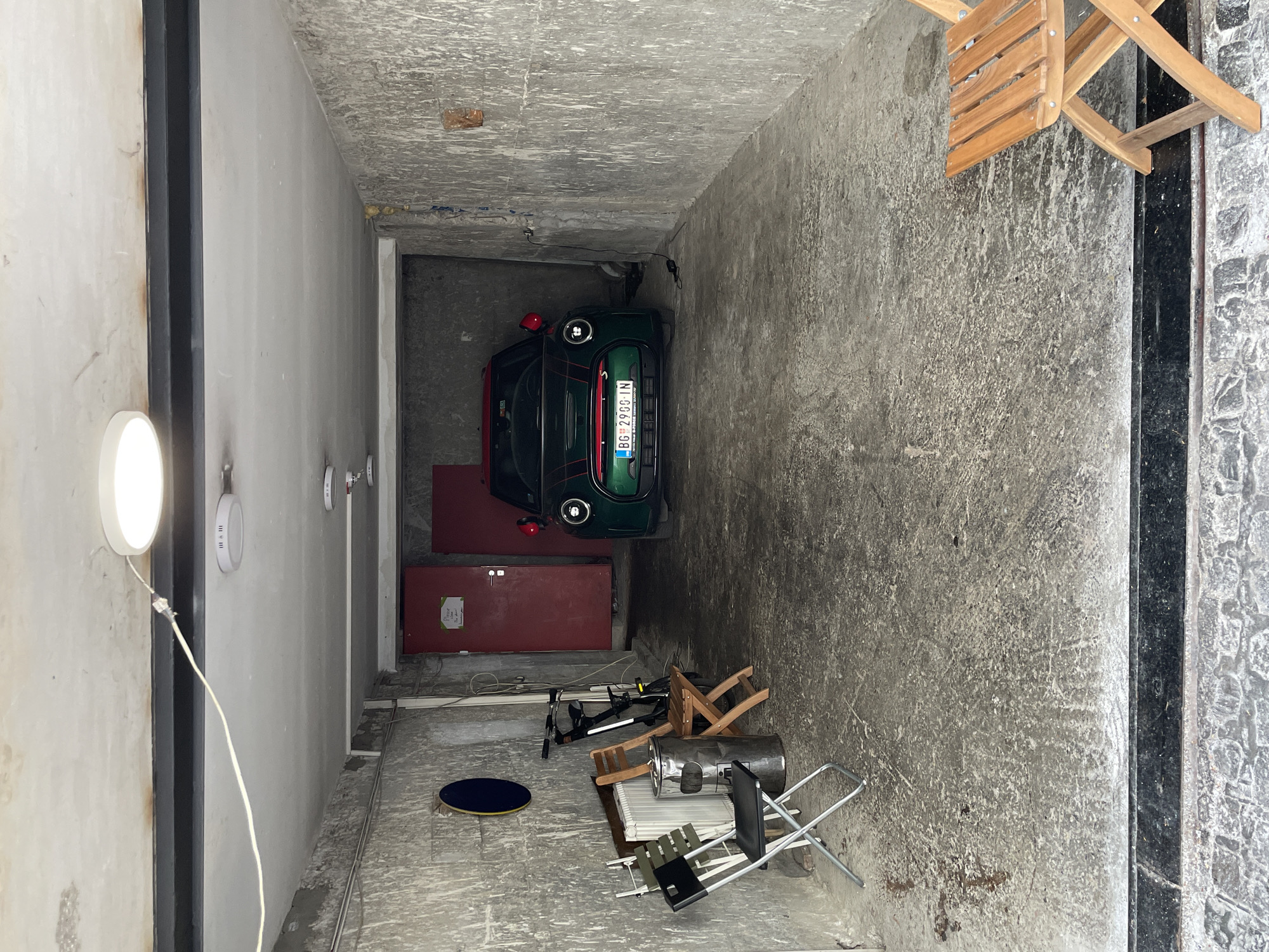 This media file captures a view of a narrow, dimly lit passage or garage. The primary subject is a dark green Mini Cooper car parked within the confined space, its license plate reading "BG 2996 IN". The car is positioned towards the back of the passage.
The setting appears to be a utilitarian and somewhat cluttered environment. The walls and ceiling are made of rough concrete, showing signs of wear and age. The floor is also concrete. Scattered around the passage are various objects, including wooden chairs, what looks like a bicycle, a metal drum or barrel, and a folding metal chair. A large red door is visible on the left side of the passage, and another red structure is situated behind the car.
On the left wall, a bright, circular light fixture is illuminated, casting a glow and suggesting this might be an indoor or covered space. Several smaller circular fixtures are also visible along this wall. Cables and wires are present, connected to some of the lights and possibly to the car.
The overall impression is one of a functional, perhaps storage or parking area, with objects that might be used for outdoor activities or simply stored there. There is no indication of people or a specific event taking place; the scene is static. The lighting is somewhat uneven, with the bright fixture creating a contrast to the overall dimness. The concrete textures and the presence of various stored items suggest an urban setting, consistent with the provided location context of Belgrade, Serbia.