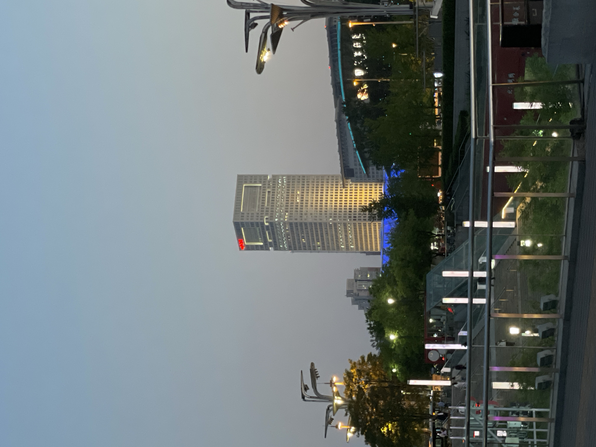 The image captures an outdoor scene in Chaoyang District, China, at dusk or twilight. The skyline is dominated by a tall, modern skyscraper with a grid-like pattern of illuminated windows. The building features a distinct red logo on its upper portion. The lower section of the image is characterized by a pedestrian walkway with illuminated structures and railings. Trees are visible along the walkway, interspersed with street lamps. People are present, though not prominently featured, suggesting a public area. The sky is overcast and evenly lit, contributing to the dim atmosphere. The composition is slightly askew, with the image rotated counterclockwise.