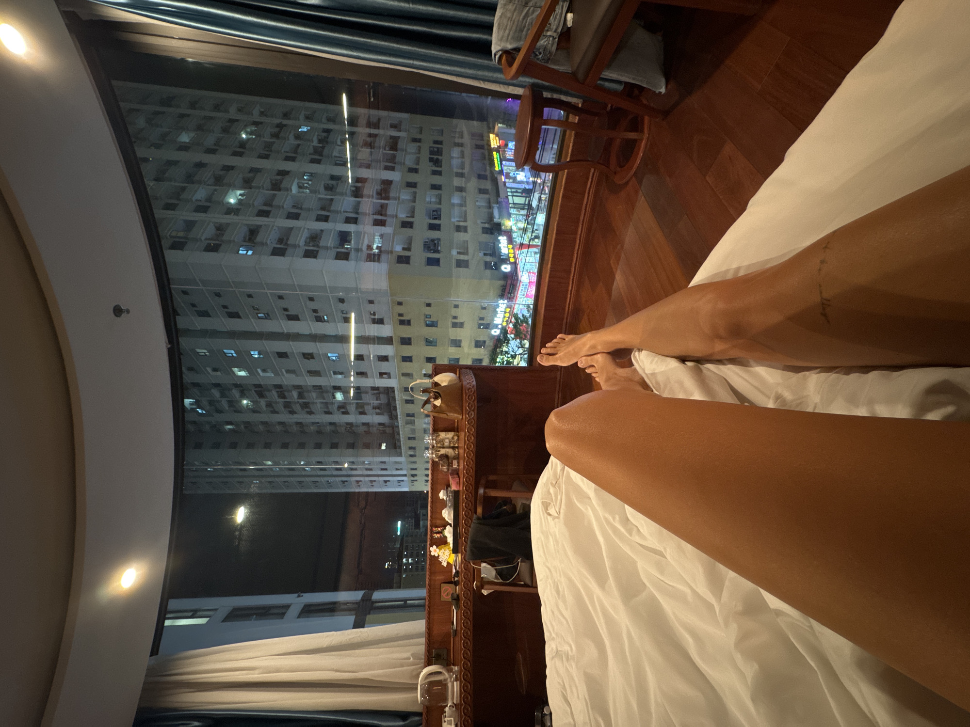 The image, rotated counter-clockwise, depicts a view from a hotel room in Đà Nẵng, Vietnam at night. The primary focal point is a pair of tanned legs resting on a white bedsheet. Through a curved window, a cityscape is visible, showing tall buildings illuminated with lights. A small wooden chair and table are positioned near the window. A dark wooden desk cluttered with various personal items is also present. The room is dimly lit, with a cool hue emanating from the city lights outside.