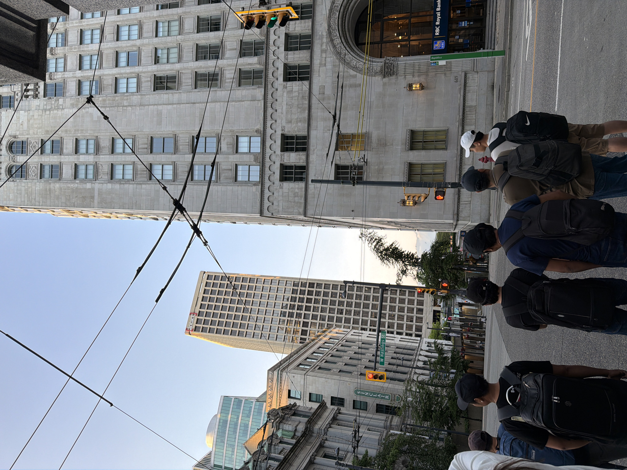 The image depicts a street scene in downtown Vancouver, Canada, likely near the intersection of W Hastings Street and Seymour Street. In the foreground, a group of five individuals, all wearing backpacks and facing away from the camera, are standing on a paved street. The individuals are casually dressed.
Behind the group, a tall, grey stone building with numerous windows dominates the right side of the frame. This building appears to be an older, ornate architectural style. To the left of this building, another, taller, modern building with a grid-like facade is visible. Further to the left, a classical-style building with stone detailing and arched windows can be seen. Overhead, a network of thick cables, likely for streetcars or other utilities, crisscrosses the sky. Traffic lights are visible on poles, with one showing a red light and another showing a green light. A street sign indicates "W Hastings St". A sign on the grey stone building to the right reads "RBC Royal Bank". The sky is a clear, pale blue, suggesting it is late afternoon or early evening. Trees with green foliage are present in the mid-ground. The overall impression is an urban environment with a mix of historical and modern architecture.