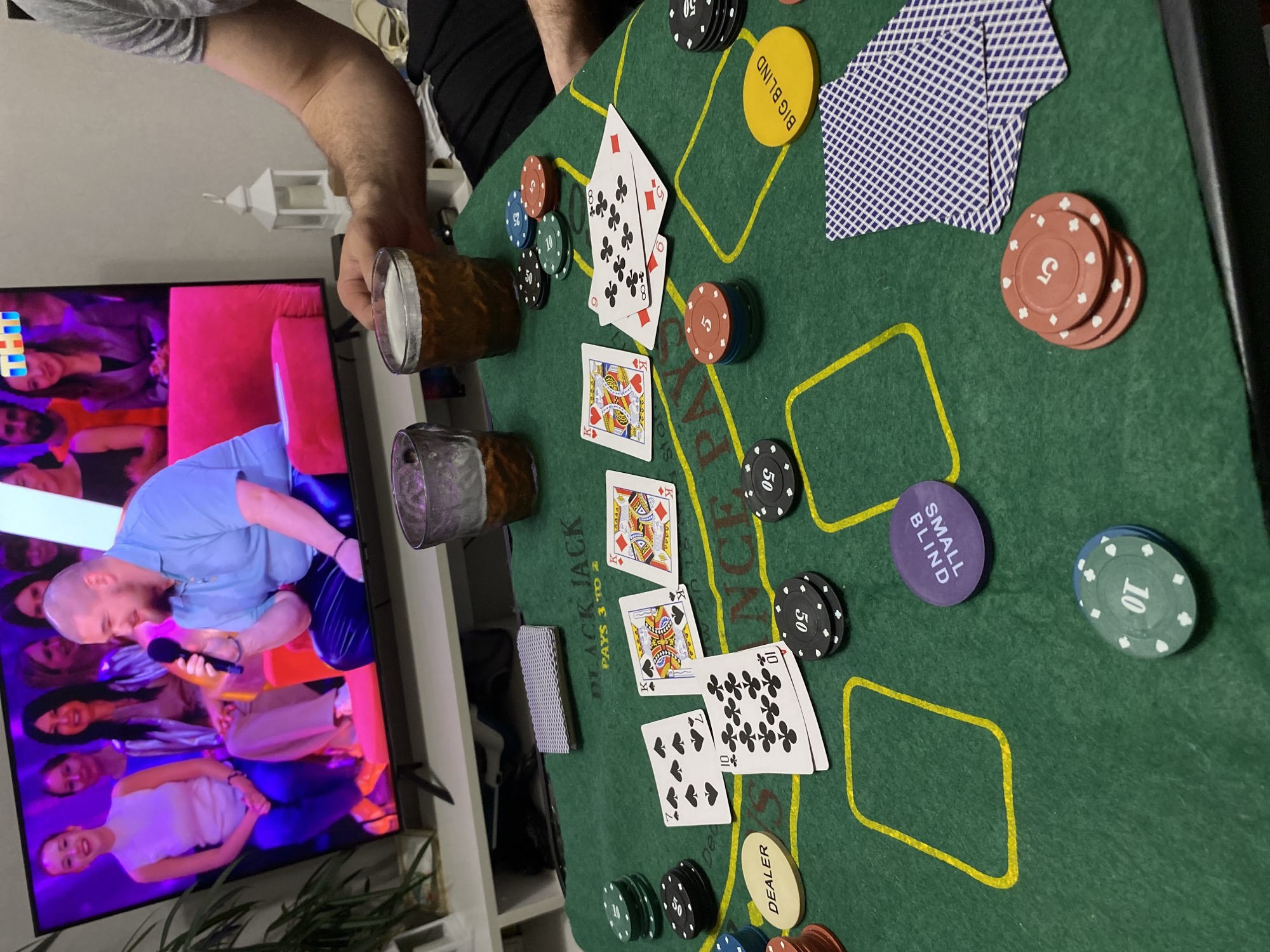 The image depicts a poker game in progress. A green poker table with yellow markings is prominently featured, scattered with poker chips of various denominations (5, 10, 50), cards face-up on the table, and labeled "BIG BLIND", "SMALL BLIND", and "DEALER". The cards show a pair of kings. There is a stack of cards and chips. Two glasses, seemingly filled with a dark liquid, are also visible. In the background, a television is showing a program. Parts of people are visible around the table: an arm holding one of the glasses, the person next to it is wearing a black shirt. The location is not mentioned in the image and cannot be determined from the available information.