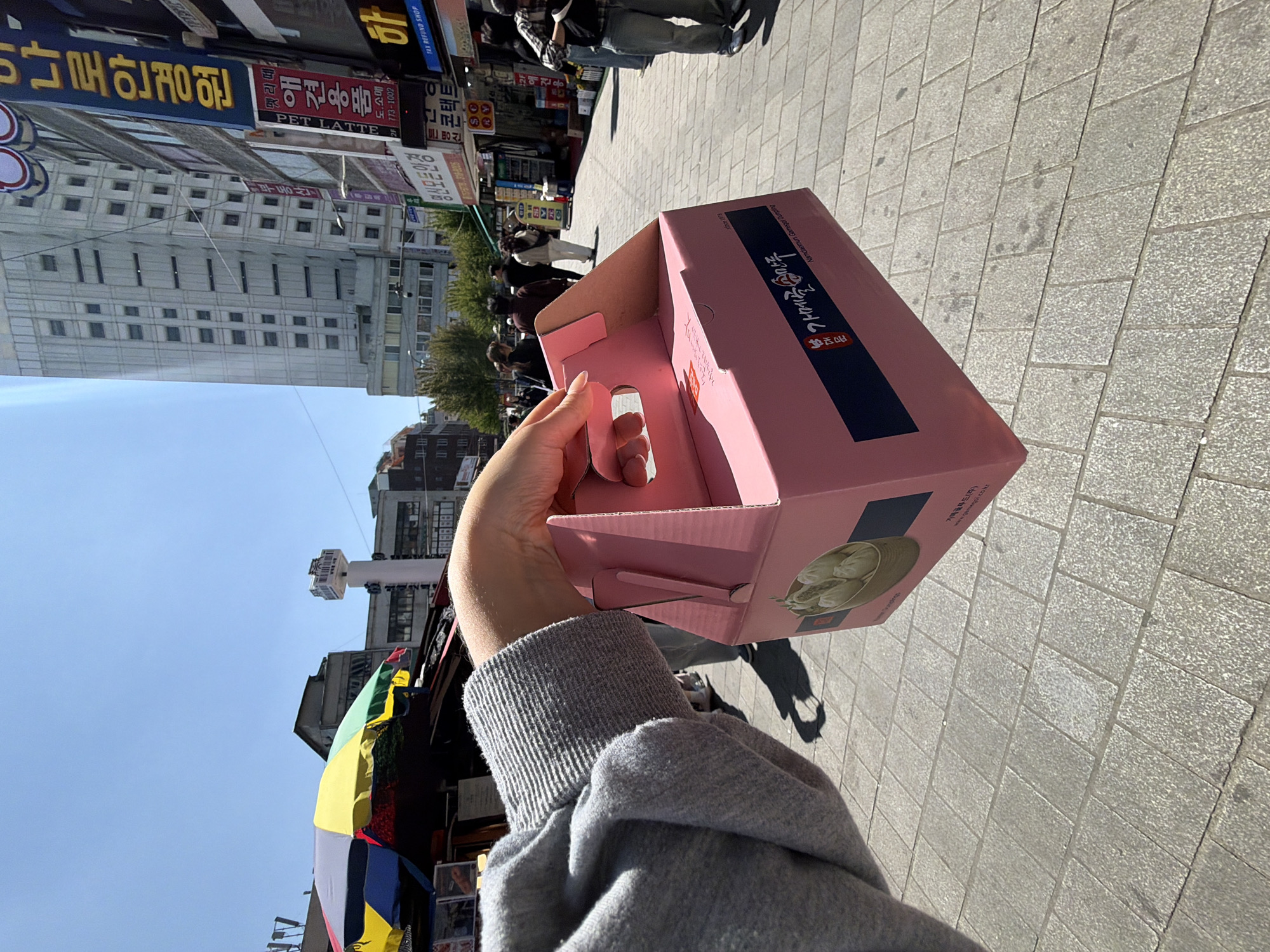 A first-person perspective image shows a person holding a pink cardboard box with a handle in an urban setting. The box appears to contain dumplings, as depicted in a graphic on the side, along with Korean text. The background reveals a paved street, buildings, and pedestrians. Korean signage is visible on the buildings, including a store named "Pet Latte" and another related to eyeglasses. A multicolored umbrella is partially visible on the left. The scene appears to be located in Seoul, South Korea, indicated by the Korean text and architectural style. The weather is clear and sunny, casting shadows on the pavement.