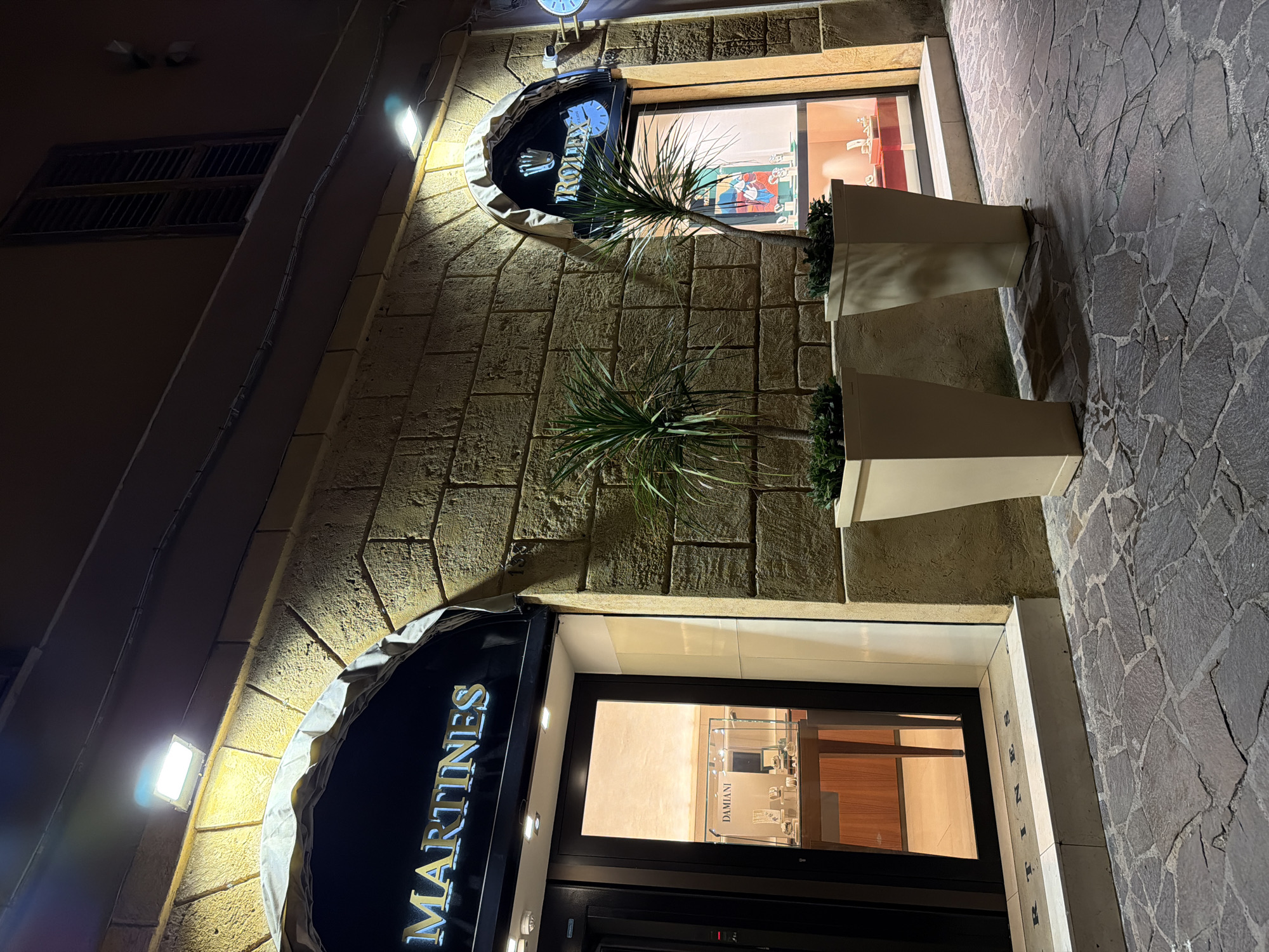 The image depicts the exterior of two adjacent luxury jewelry stores at night on a stone-paved street in Agrigento, Italy. The store on the left has a black awning with the illuminated gold lettering "MARTINES". Its glass entrance displays a jewelry case labeled "DAMIANI". The store on the right features a similar black awning with the illuminated Rolex logo and name. Both stores have well-lit interior displays showcasing jewelry. Two large, light-colored planter boxes with green plants are positioned in front of the stores. The building facade is constructed of textured stone blocks, illuminated by overhead lights. The overall ambiance is upscale and refined.
