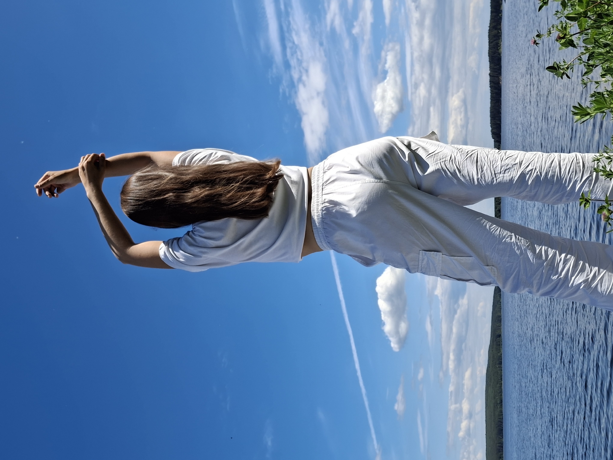 An outdoor, low-angle, full-length photograph captures a person from behind, standing at the edge of a body of water in Chaikovsky, Russia. The individual has long, straight brown hair and is wearing a white short-sleeved crop top that exposes their lower back and a sliver of midriff, along with light-colored, possibly white or light grey, cargo pants with visible pockets on the sides. Their arms are raised overhead, with the left hand gripping the right forearm.
The background features a vast, clear blue sky with scattered white cumulus clouds. A prominent, straight white contrail extends horizontally across the mid-left portion of the sky. Below the sky, a large, calm blue lake stretches across the middle ground, exhibiting slight ripples on its surface. On the far side of the lake, a dark green coniferous forest covers a low-lying landmass. In the immediate foreground, to the right, green foliage with several small pink flowers is visible, consistent with clover. The scene is brightly illuminated by natural light, indicating a sunny day.