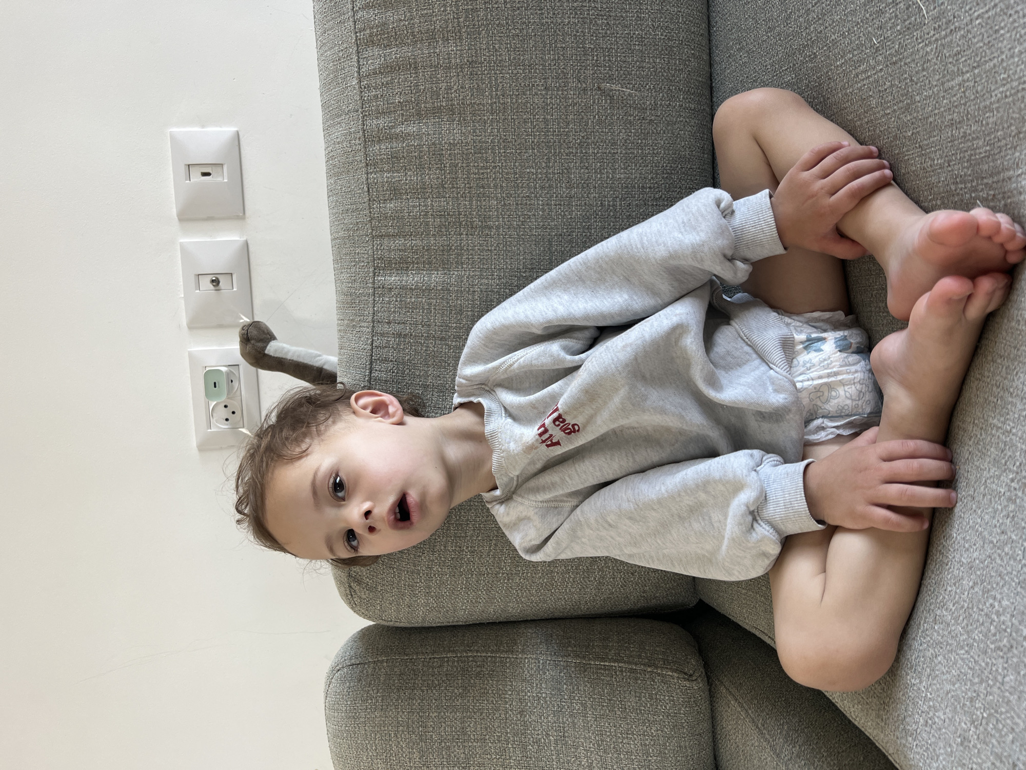 A toddler wearing a light gray sweatshirt and a diaper is sitting on a gray sofa. The child has brown hair and is looking directly at the camera with their mouth slightly open. The background consists of a white wall with electrical outlets and a coaxial cable outlet. A stuffed animal tail is visible near the child's head. The location is Kiryat Bialik, Israel.