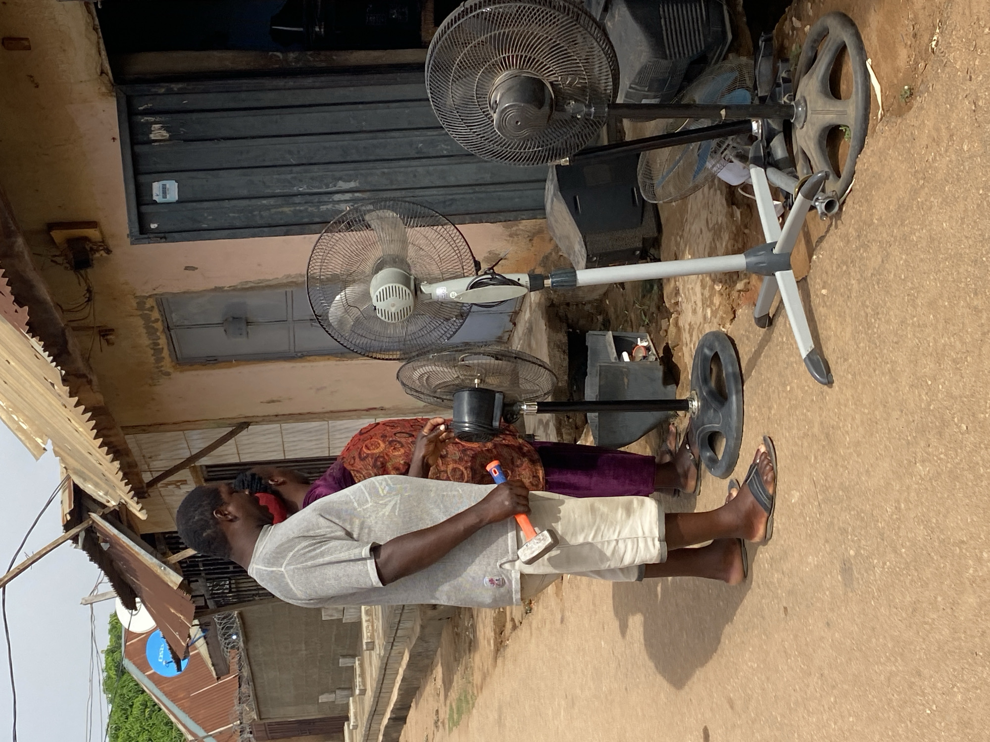 Outside a building in Narayi, Nigeria, a man in a light-colored shirt and white shorts is holding a hammer. A woman in a patterned top and purple pants stands beside him. Several electric fans, some disassembled, are placed against the building wall. The building has a closed metal shutter and a smaller door. The ground is a mix of concrete and dirt. In the background, a partially visible house with a satellite dish on the roof can be seen.